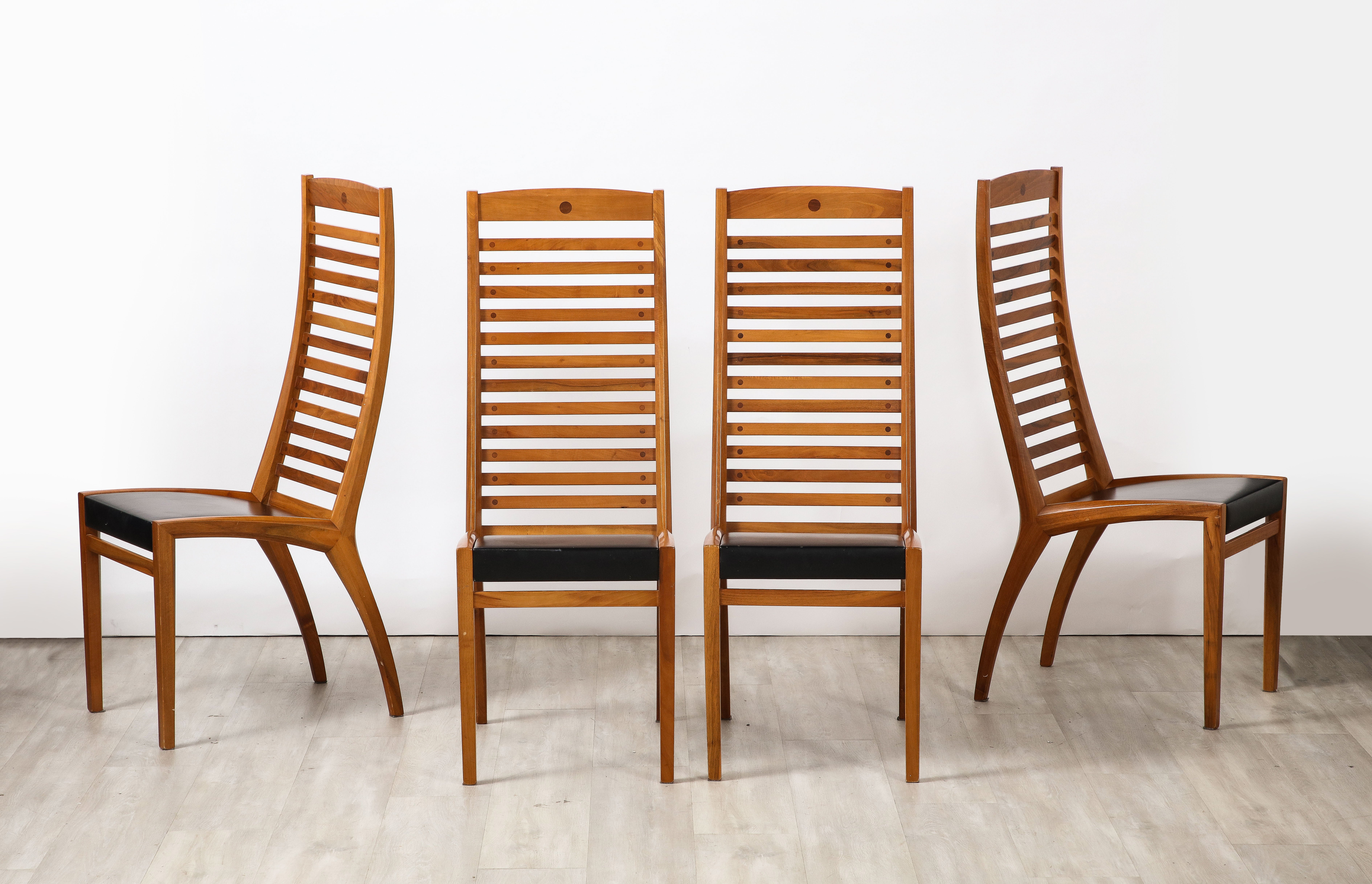 A set of four walnut ladder back dining chairs with original black leather seats, the seats and back legs are angled, with straight legs in the front.  The chairs from every perspective represent an elevated and sleek modernist design of the era.