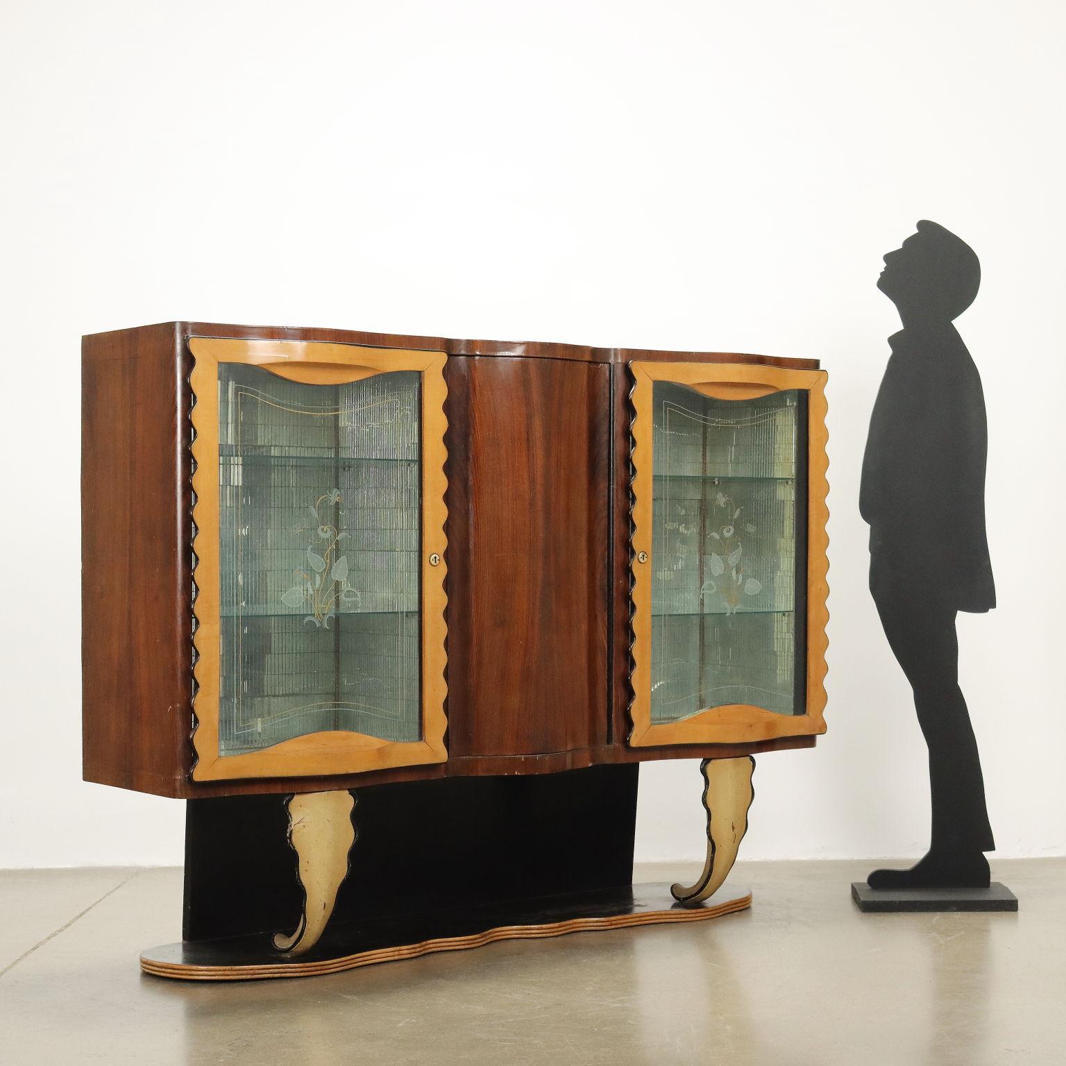 Vitrine avec portes à charnières latérales, intérieur avec prises en verre miroir, compartiment central dissimulé pour le bar avec porte-verres et porte-bouteilles. Bois plaqué noyer, pieds en bois sculpté laqué.