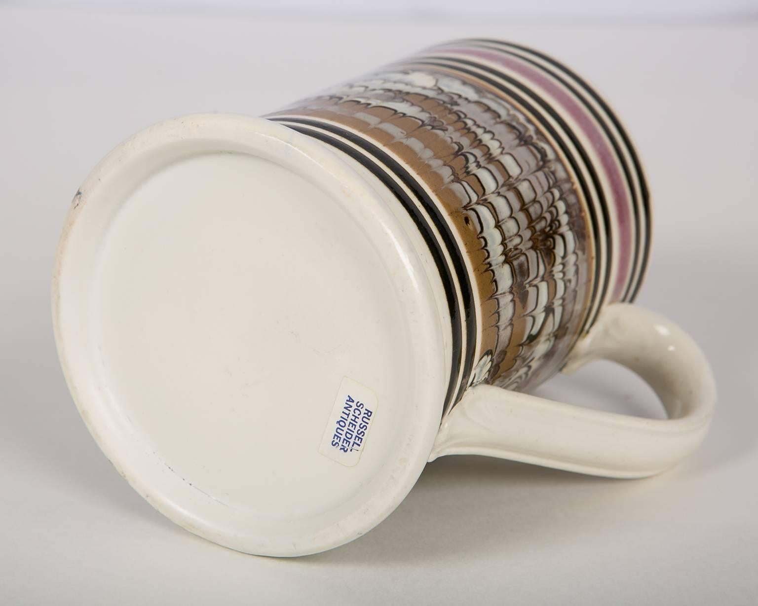 An exceptional and rare mochaware pint mug. The top of the mug is decorated with a band of lavender slip. Below the band is a rare design of combed slip in three colors: lavender, light blue, and midnight brown. The unique design is created by the