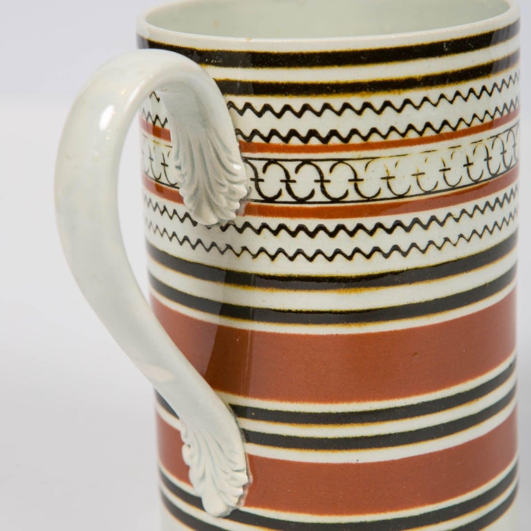 A Mochaware mug decorated with bands of slip in light and midnight brown. Between the midnight and light brown slip bands are three bands of inlaid rouletting decoration in geometric patterns. Although made circa 1815, the inlaid rouletting gives