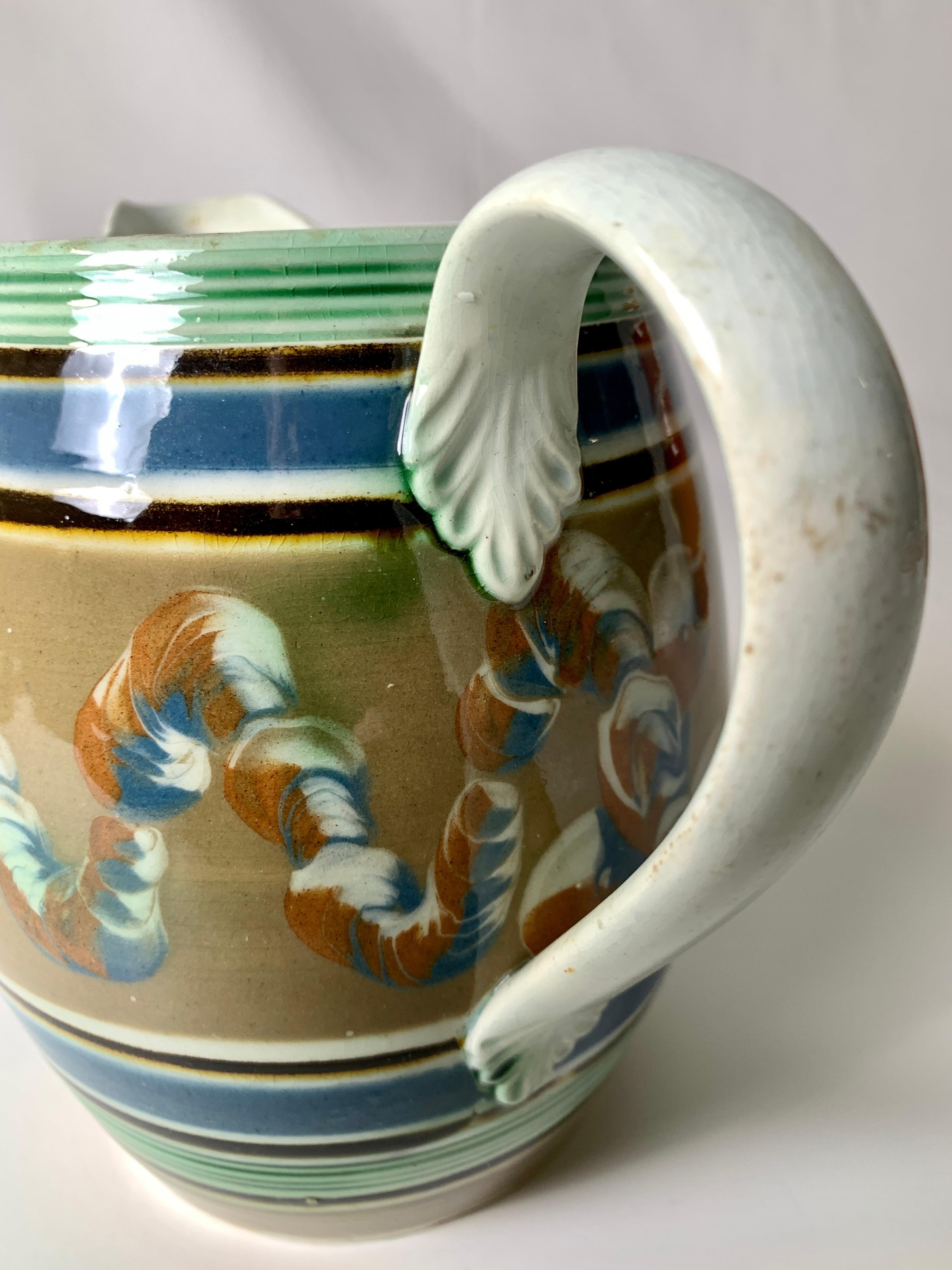 Every piece of Mochaware is unique. This pitcher is decorated with slip (colored clays) in a three-color cable pattern of soft blue, beige, and chocolate brown on a beige ground. Around the top and bottom are grooved rings decorated with green