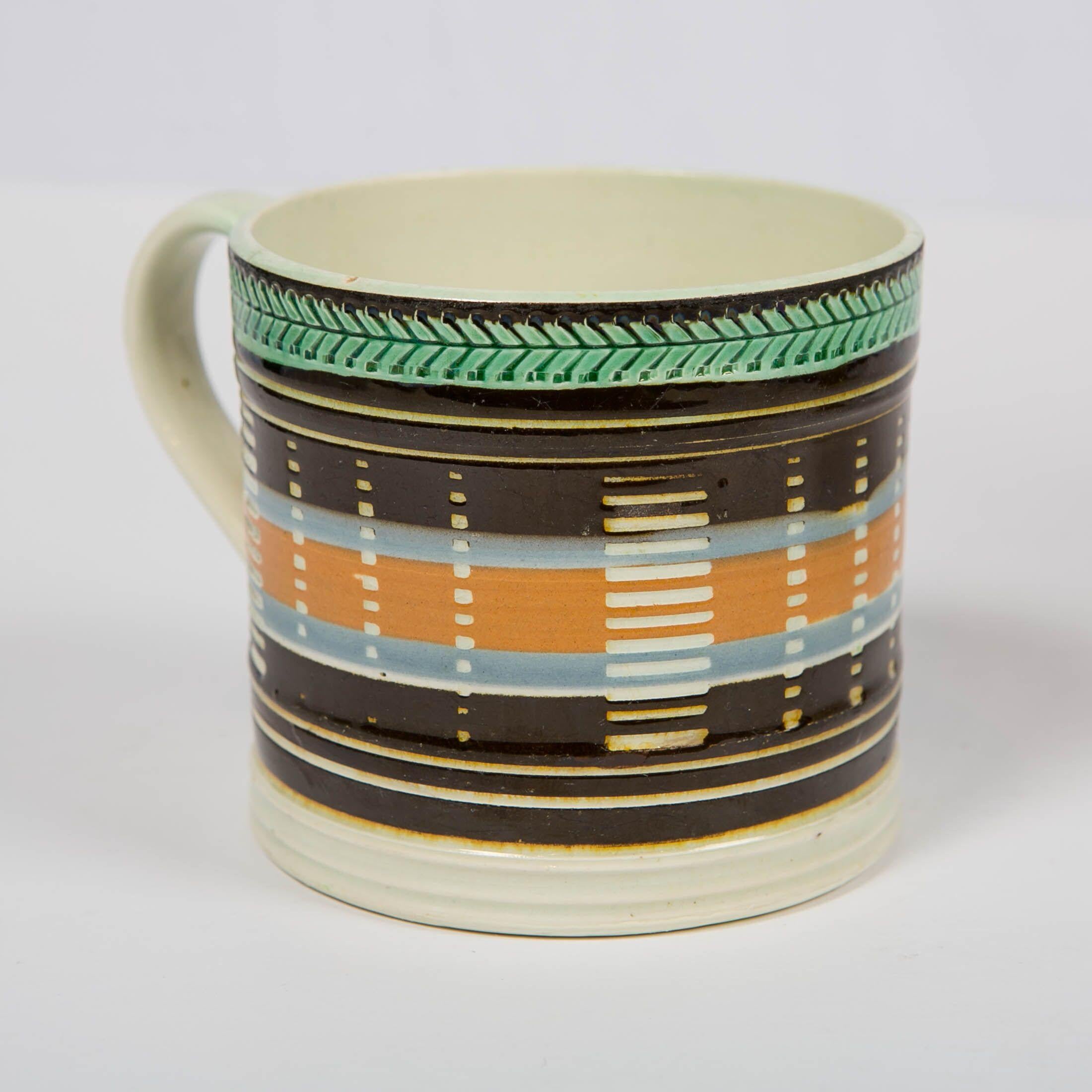 A mochaware mug made in England, circa 1820. The mug is made of pearl-glazed creamware. The decoration has dramatic black, light blue, and orange bands of slip, which are cut through in 