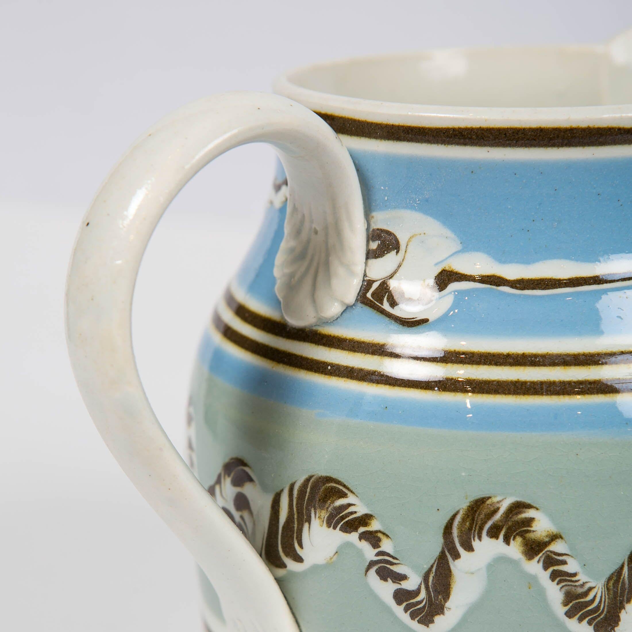 A Mochaware pitcher made of pearl-glazed creamware in England, circa 1820. The pitcher is decorated with bands of baby blue and moss green slip and thin bands of black slip. The handle has acanthus leaf terminals.
The most notable features are the