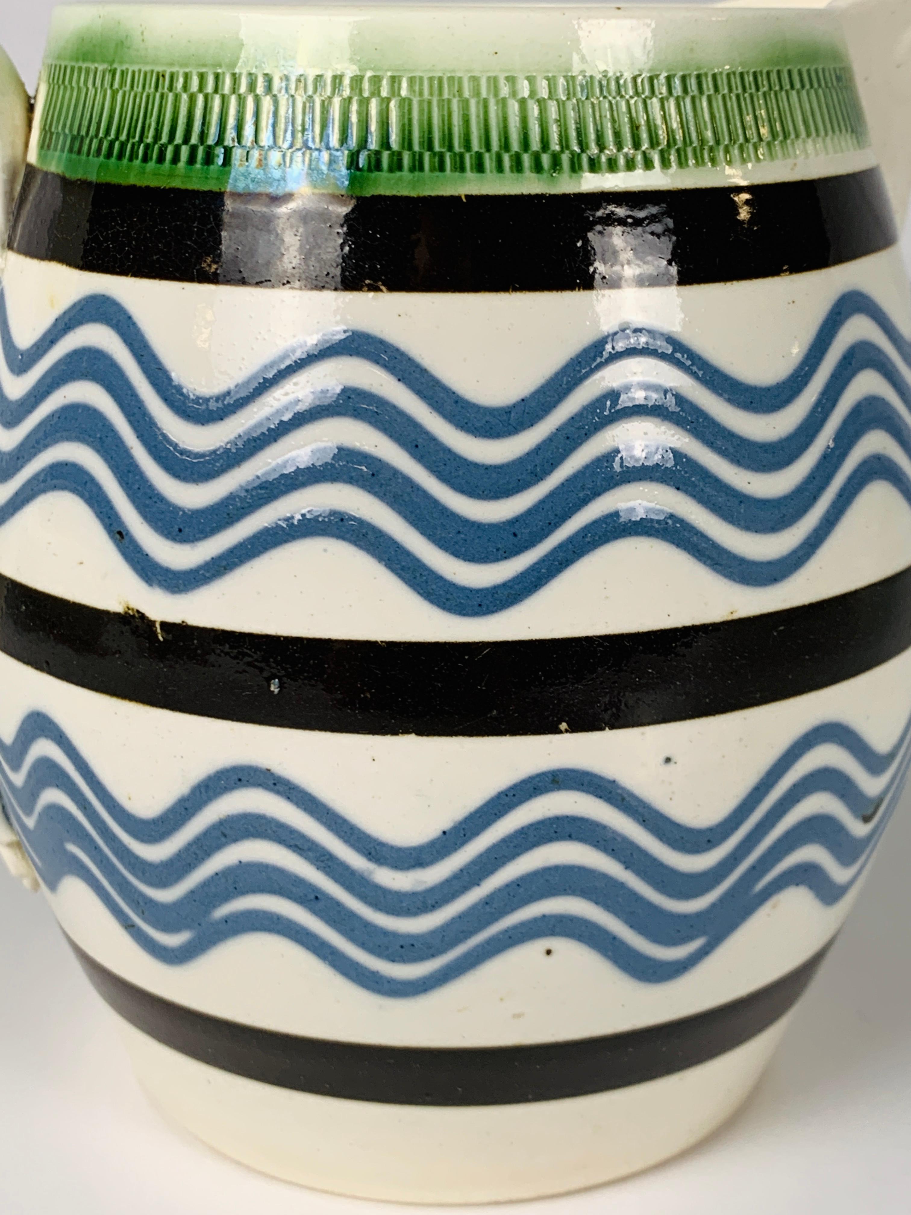 A mochaware pitcher with two panels showing four wavy sky-blue trailed slip lines, around the top edge a band of green glazed rouletting, and three bands of black slip. The color combination on this crisp white ground is exciting.
The spout has an