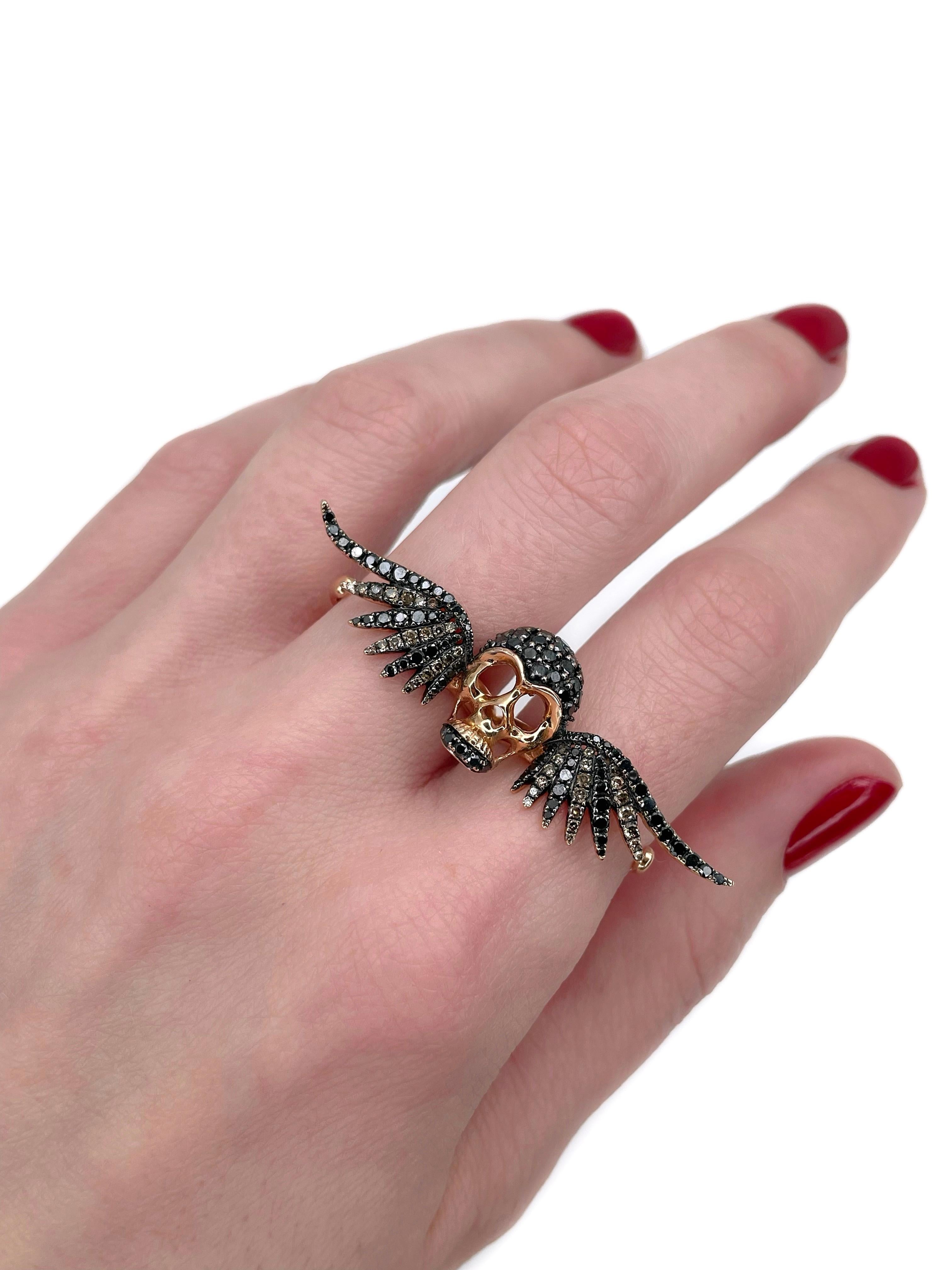 This is a modern design two finger cocktail ring crafted in 18K gold. Circa 2000. 

The piece depicts a skull with the wings. 

The ring features:
- 89 black diamonds (round brilliant cut, TW 1.10ct, Declasse) 
- 39 brown diamonds (round brilliant