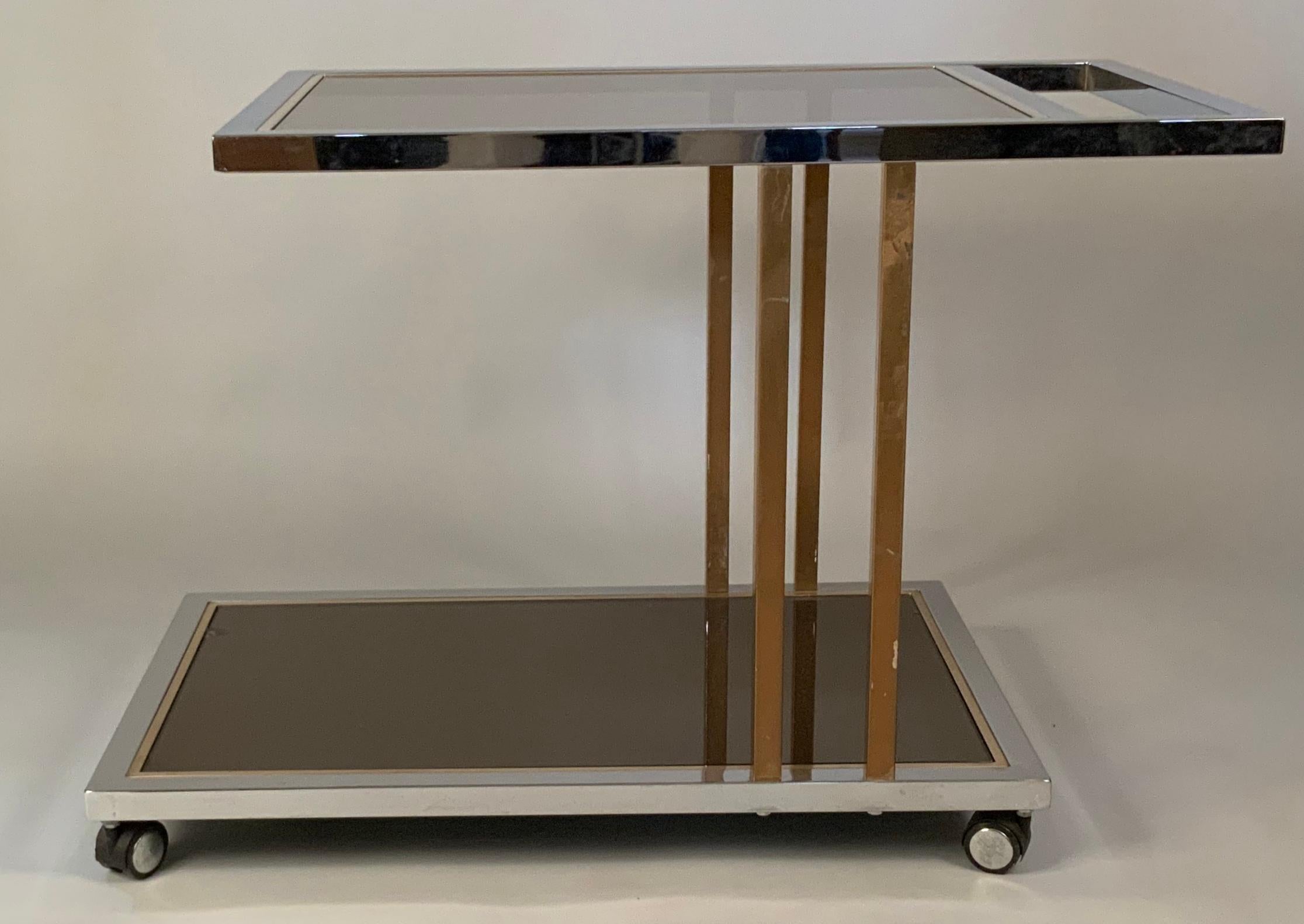 A very handsome modern 1970's rolling bar cart, with a frame of chrome and vertical bars with a brass finish. The two levels have smoked glass shelves. beautiful and functional design, with an integrated handle at one end, and the lower shelf