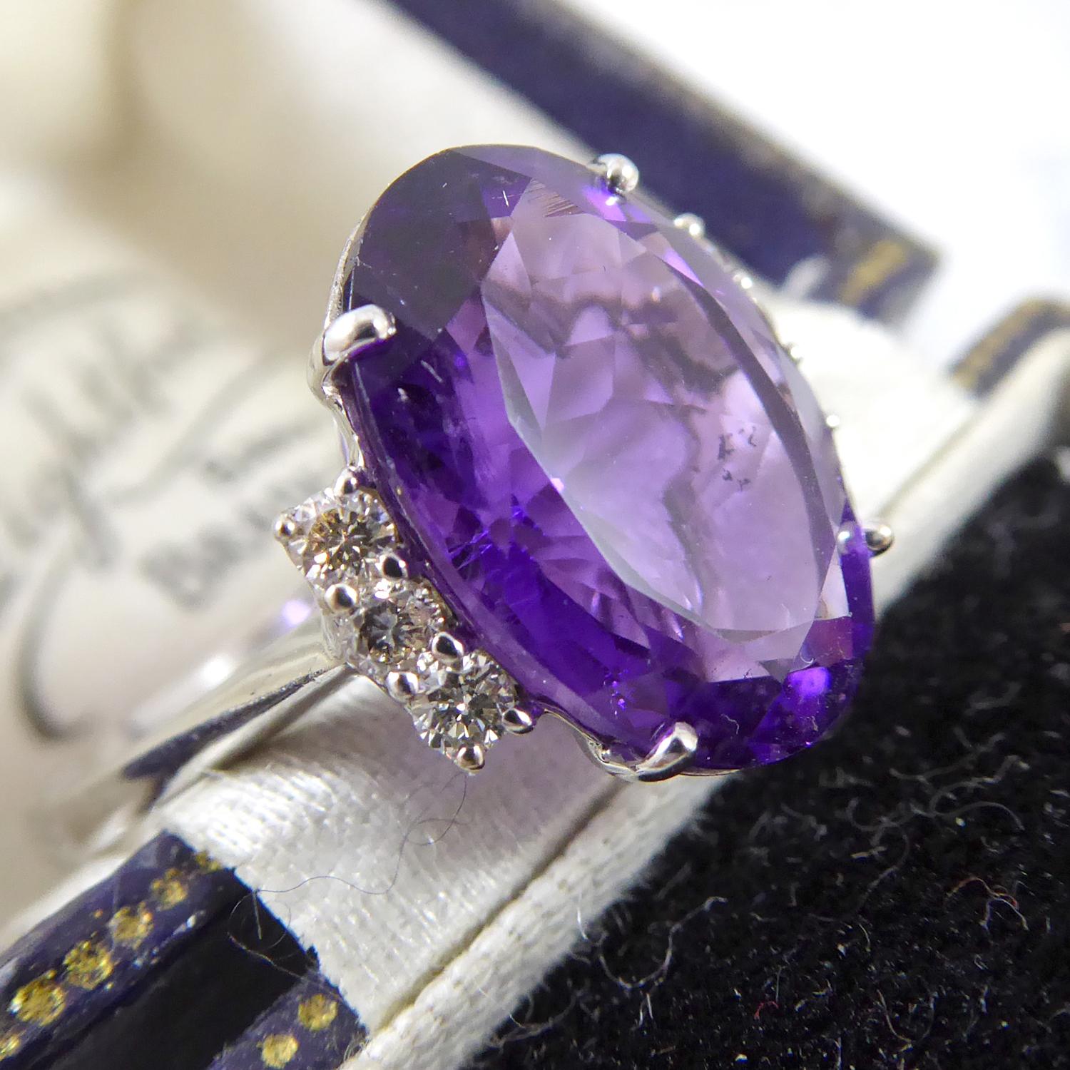 A contemporary design dress ring set with a 4.13 oval cut amethyst measuring approx. 12.98mm x 9.93mm x 6.78mm and weighing 4.13ct.  Held in a white four claw setting and flanked to each side by a vertical row of three round brilliant cut diamonds