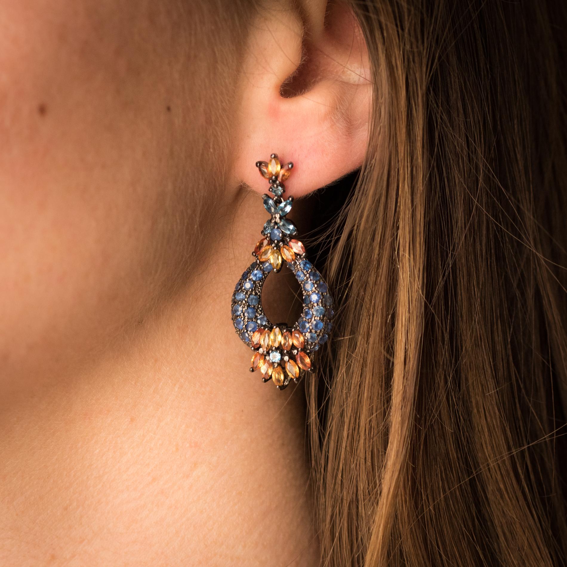 Earrings in silver, black rhodium.
These dangle earrings are composed of a motif set with 3 shuttle-shaped orange sapphires and a round blue sapphire. It retains as a pendant a motif set with blue round sapphires, shuttle-cut orange sapphires and