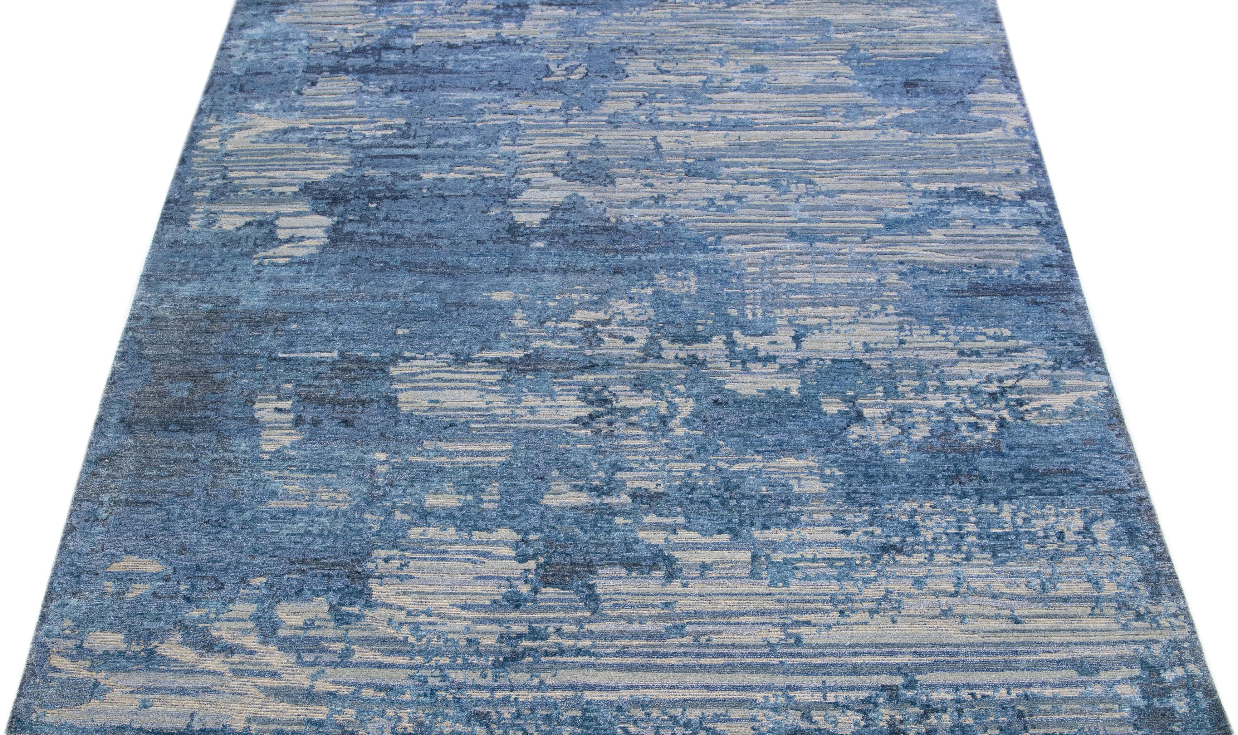 Dieser indische Teppich aus einer Mischung aus Wolle und Seide zeigt ein graues Feld mit einem abstrakten Muster, das mit blauem Satin verziert ist. Seine komponierten Materialien sorgen für Robustheit und Langlebigkeit, während sein ornamentales
