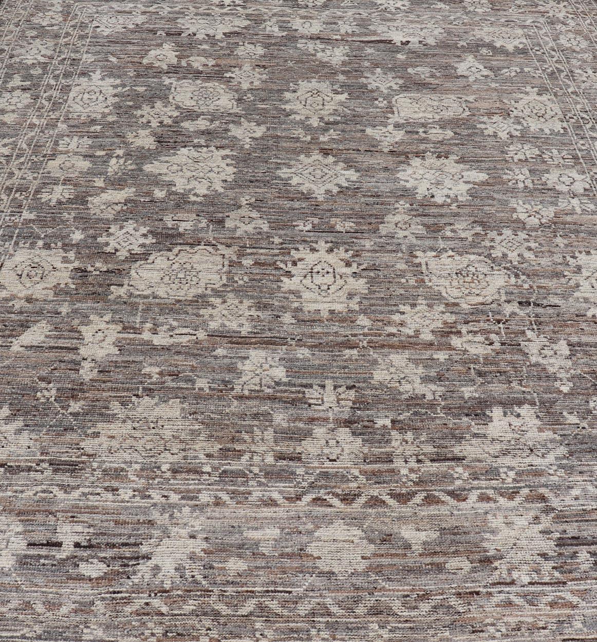Dieser moderne Teppich im Oushak-Stammesdesign wurde aus Wolle handgeknüpft. Der Teppich zeichnet sich durch ein modernes, subgeometrisches Blumenmuster aus, das von einer komplementären, mehrstufigen Bordüre umgeben ist. Der Teppich ist in Braun-,