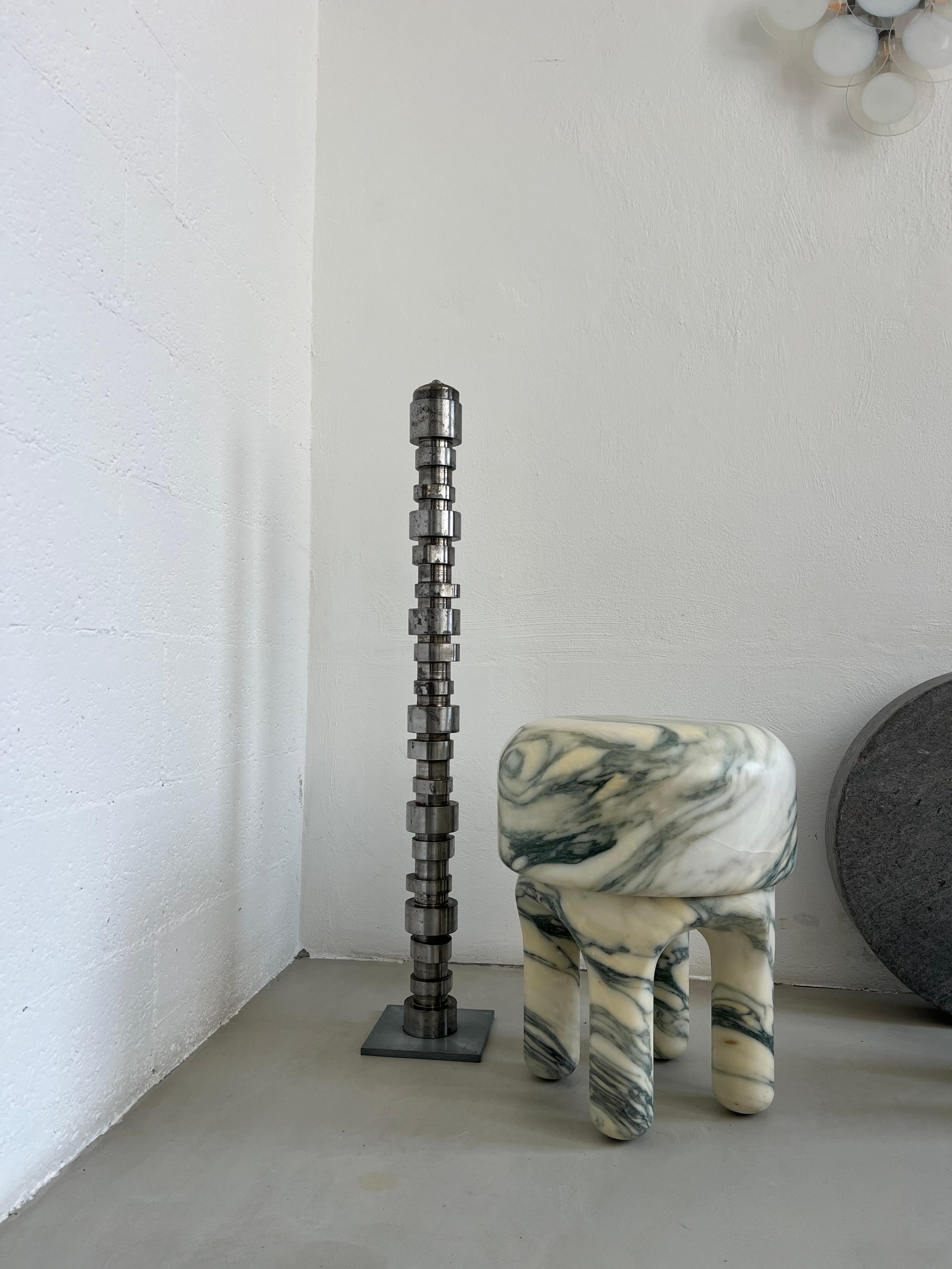 A slim freestanding steel sculpture that embodies the essence of an abstract, irregular vertical camshaft, a striking representation of strangeness and the beauty of irregularity. The sculpture rises with an unexpected, organic silhouette, featuring