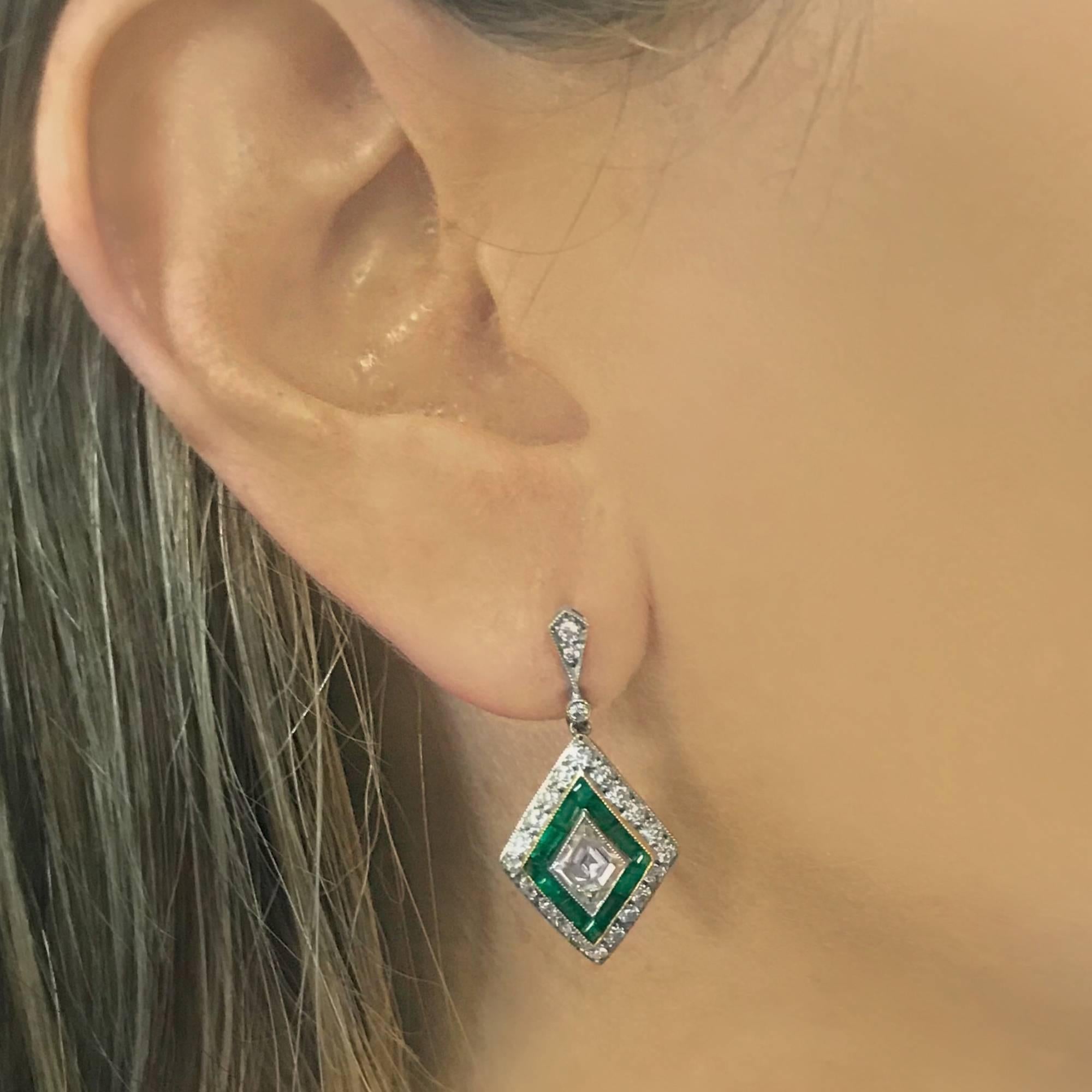 Stunning Art Deco inspired dangle earrings featuring 2 antique rhombus cut diamonds weighing approximately 1.10 carats total, G-H color, VS-SI clarity, set in platinum and framed with milgrain. The diamonds rest on a bed of 24 vivid green emerald