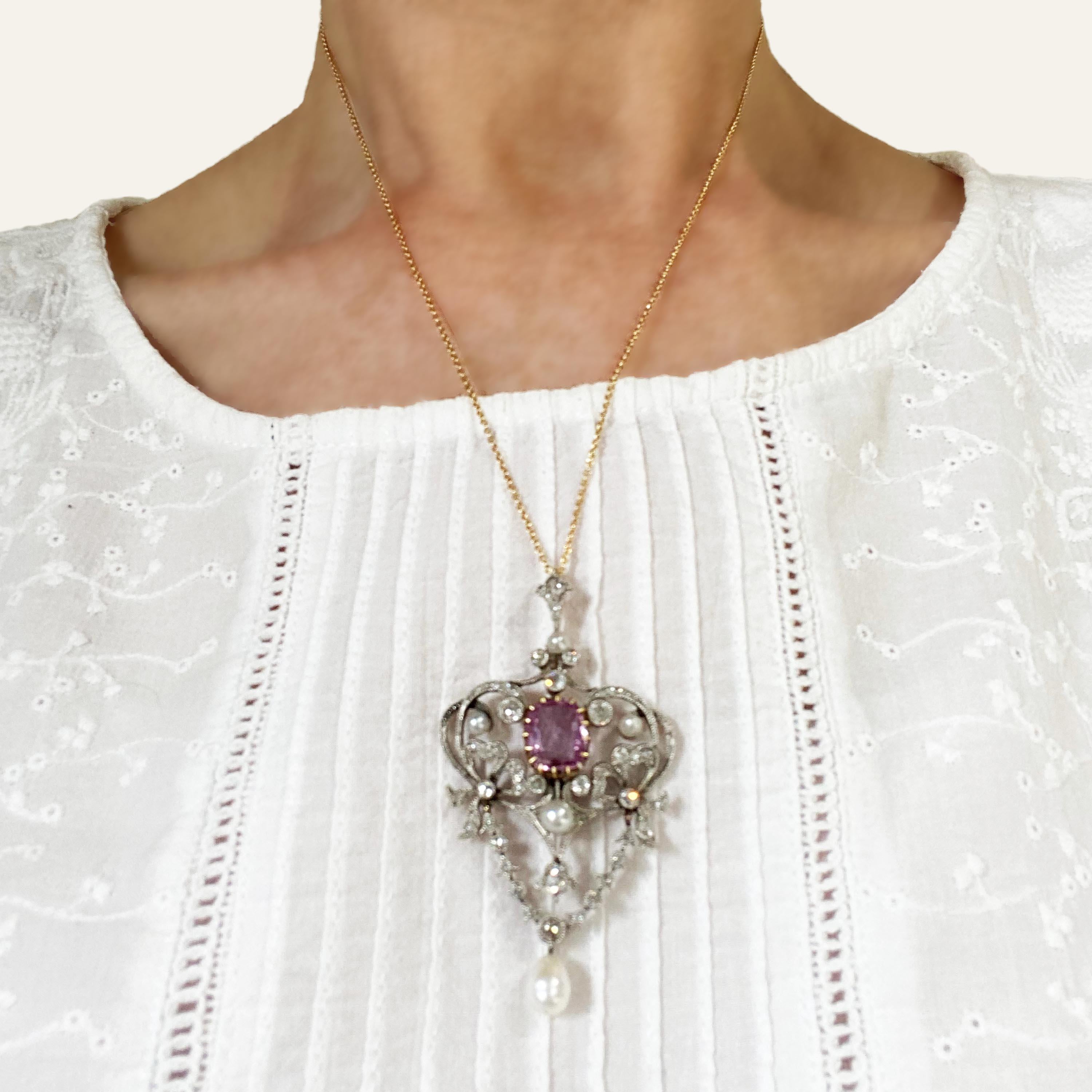 A modern belle époque style pendant, set with a native cushion-cut pink sapphire, weighing approximately 3.00 carats, in a yellow gold claw setting, in an  Art Nouveau influenced, foliate scroll surround, set with bouton and drop shape pearls and