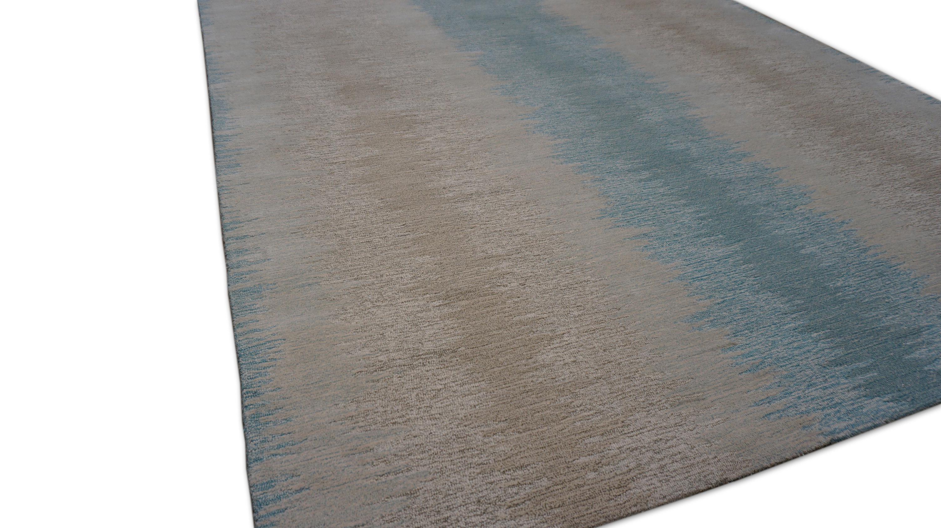 Dieser moderne, handgetuftete Wollteppich zeichnet sich durch ein ruhiges, ombriertes Streifendesign aus, das sanfte Blautöne, sandige Beigetöne und natürliches Elfenbein zu einem ruhigen Farbverlauf verbindet. Der Teppich bietet eine großzügige