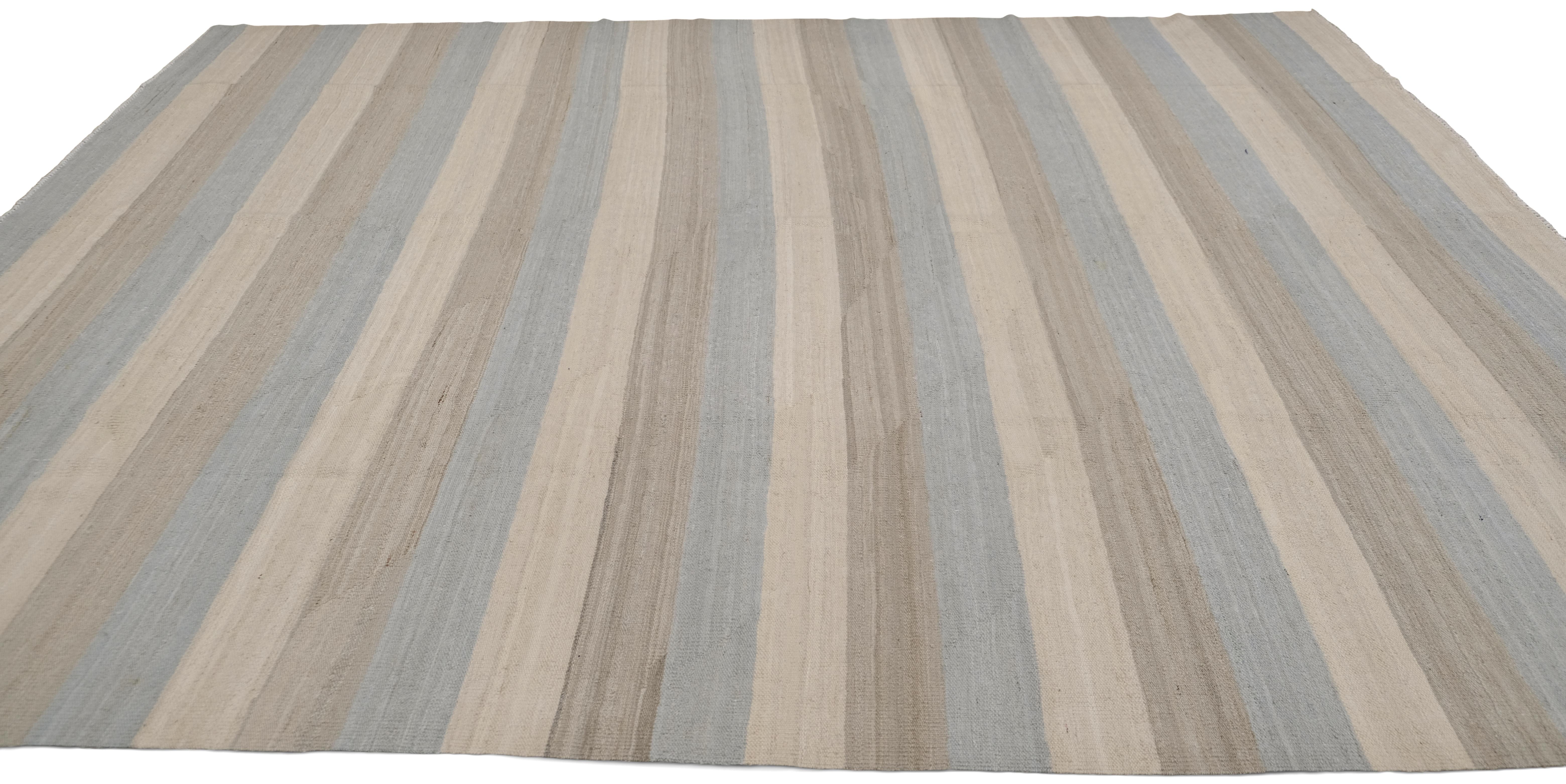 Esta alfombra de lana tejida a mano presenta un sofisticado diseño a rayas en tonos terrosos naturales, lo que la convierte en una pieza versátil y atemporal para interiores modernos y minimalistas. La suave gradación de tonos beige y azules crea