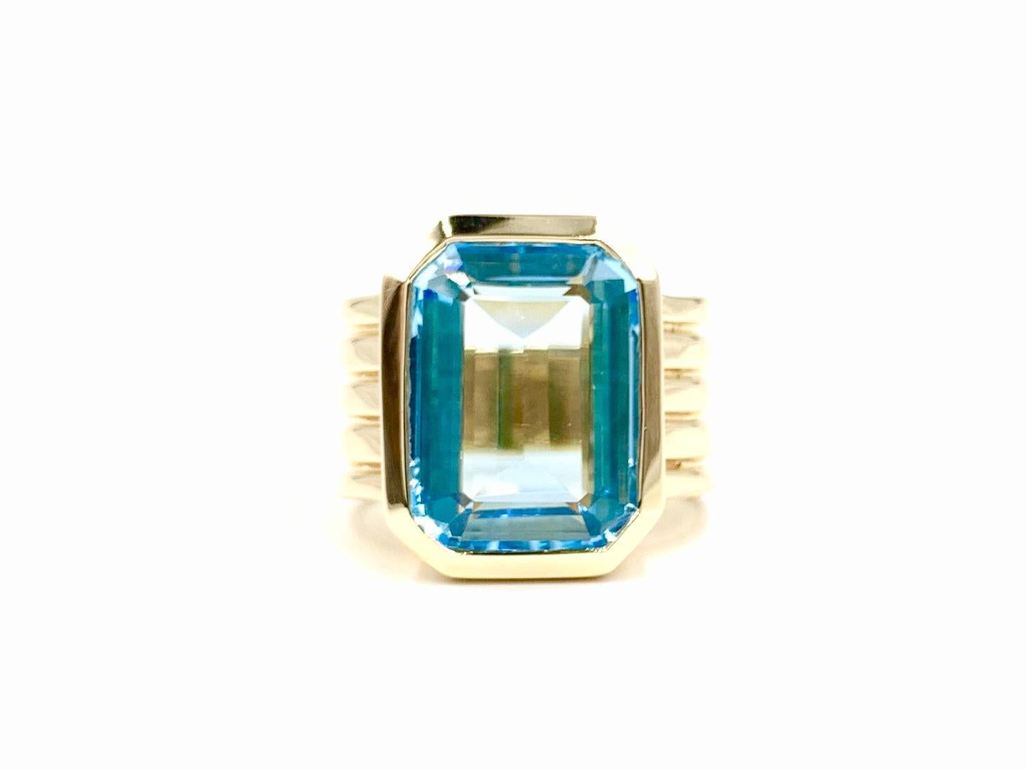 A clean and contemporary designed 14 karat gold wide ring featuring a large bezel set emerald cut vibrant blue topaz. Blue topaz measures 15mm x 11.5mm. Ring has a high-polish finish with linear grooves on the shank for added character. Width of