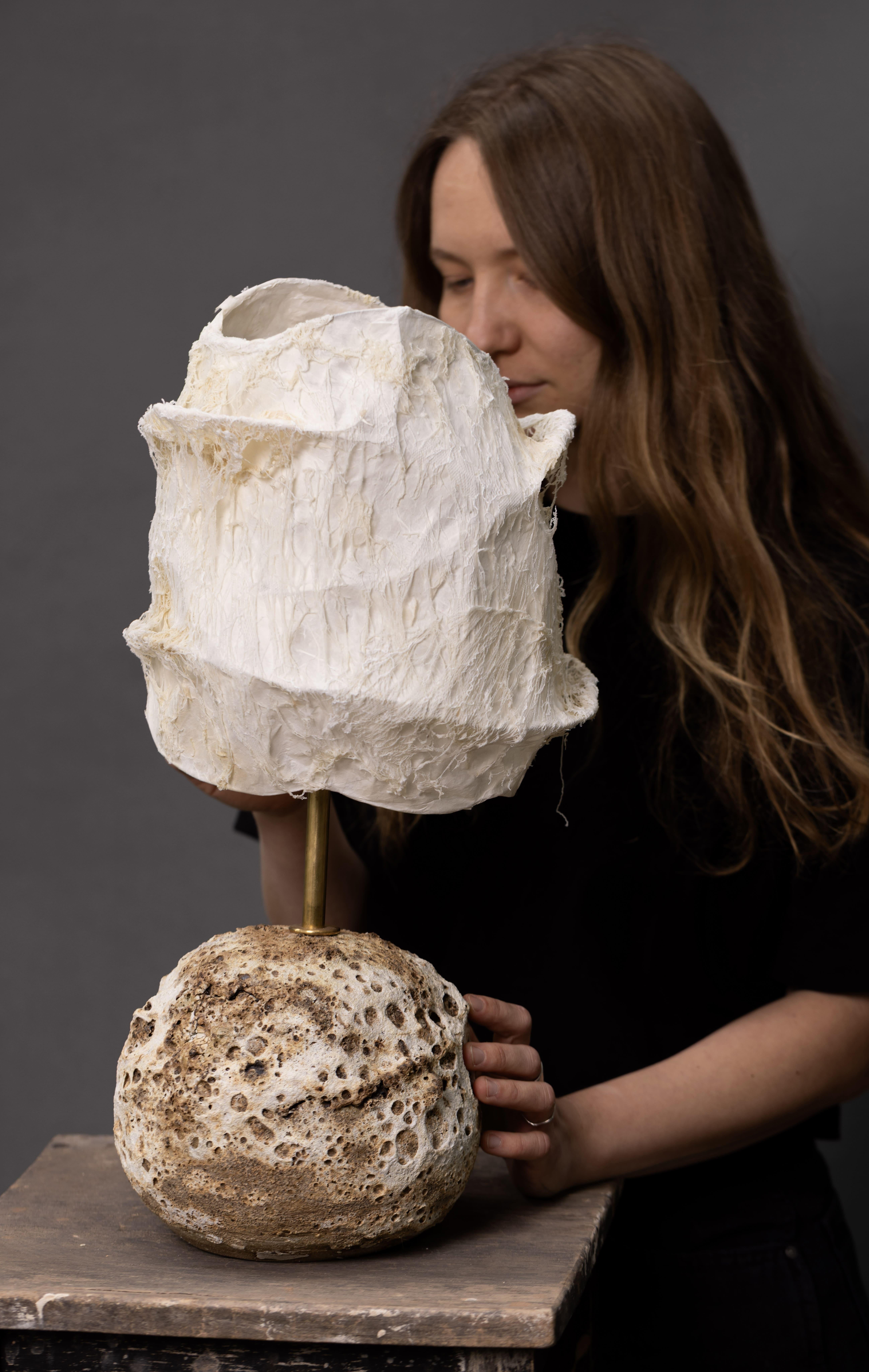 This table lamp beautifully balances contrast and craftsmanship, combining a rugged ceramic base with a soft, handmade paper shade. The base is handbuilt from natural clay and finished with a crater-like glaze that evokes the raw texture of volcanic