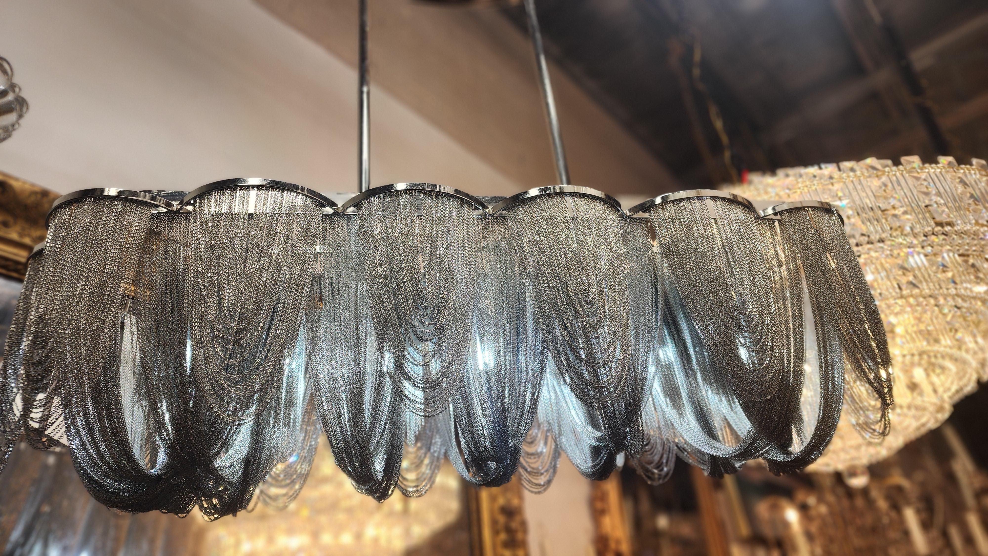 This contemporary chandelier is a captivating mix of elegance and industrial flair. Suspended from a polished metal canopy by sleek rods, the fixture features a dramatic series of cascading metal chains that form curtain-like silhouette. 