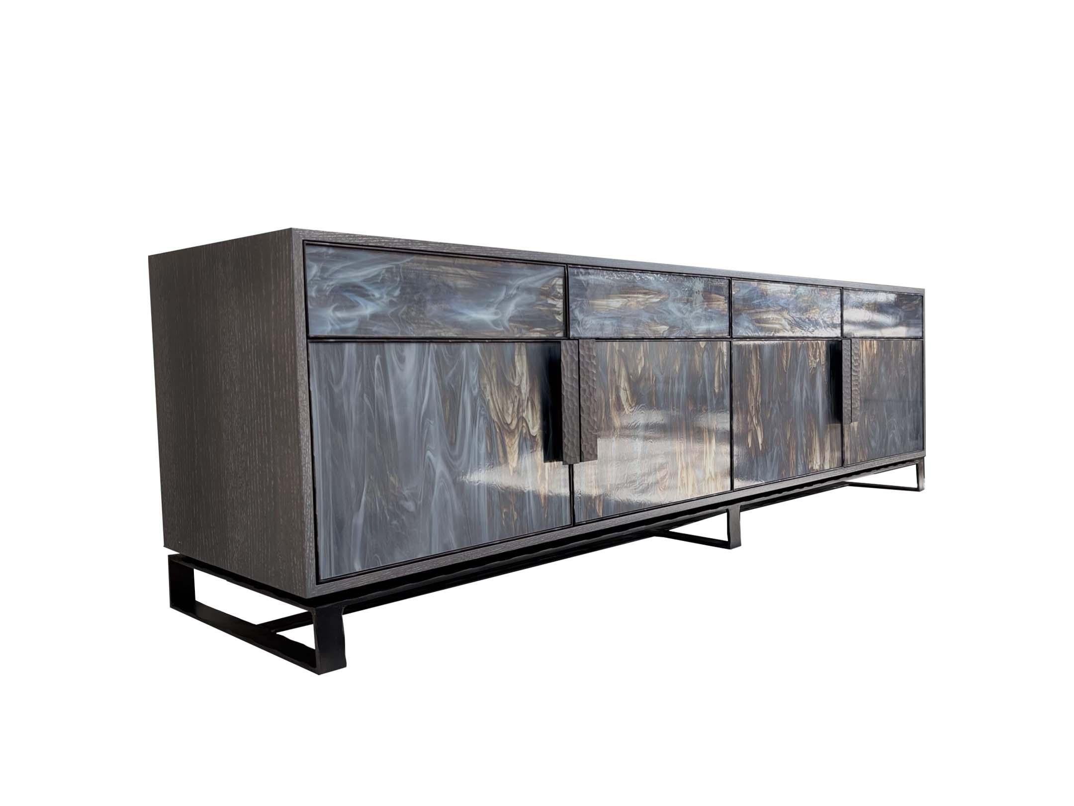 La Cómoda Moderna Chelsea de Cuatro Puertas y Cuatro Cajones presenta una carcasa de roble Earl Grey Cerused, cristal Wispy Grey Silver, cuatro puertas enmarcadas en metal Chelsea martillado a mano y cuatro cajones con pestillo táctil también