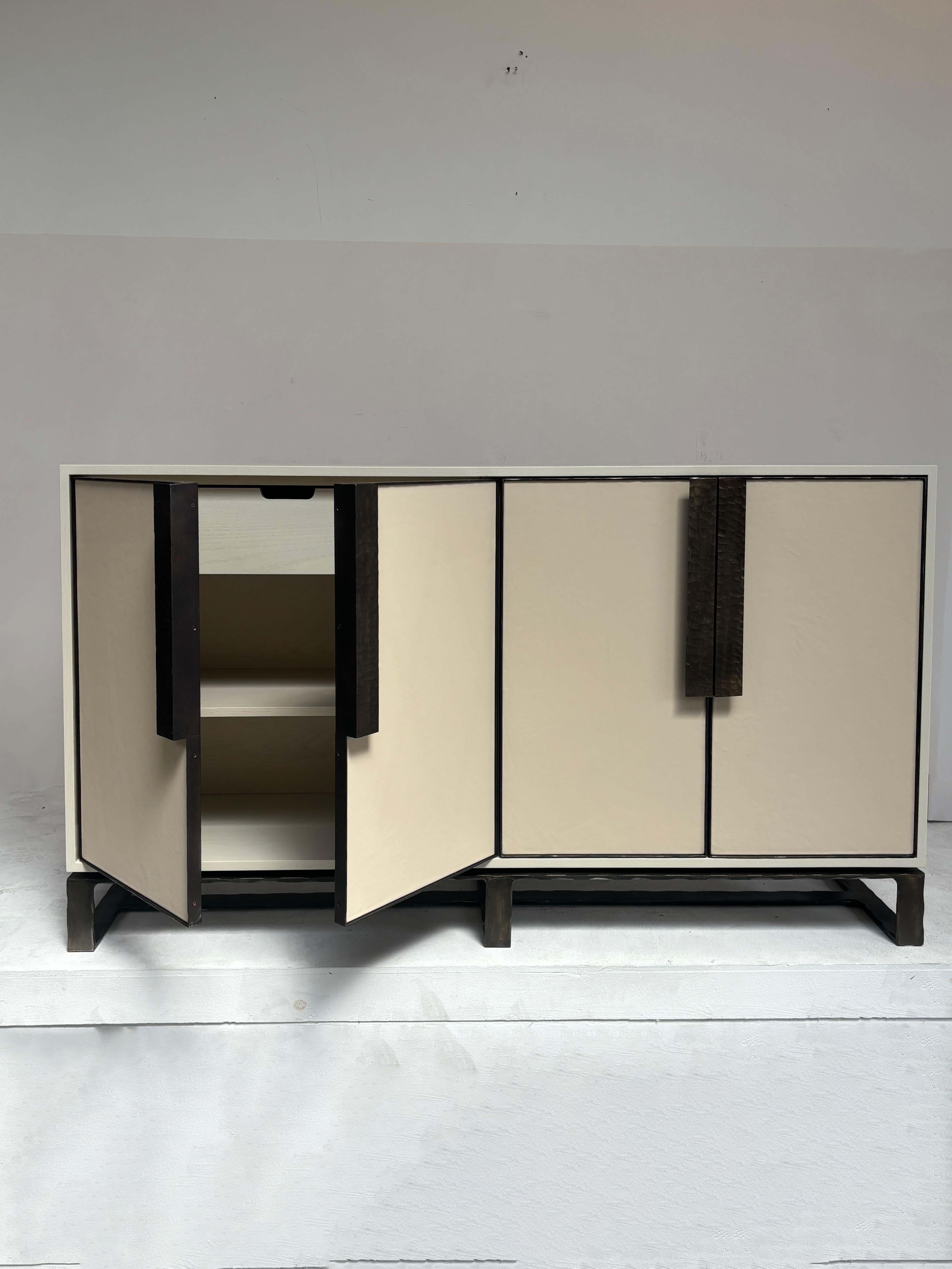 Il Buffet moderno Chelsea Leather a quattro ante di Ercole Home ha un frontale a quattro ante con finitura Sand su Oak. La pelle riveste ciascuna delle ante con cornice in metallo martellato a mano, con maniglie e base martellate a mano in finitura