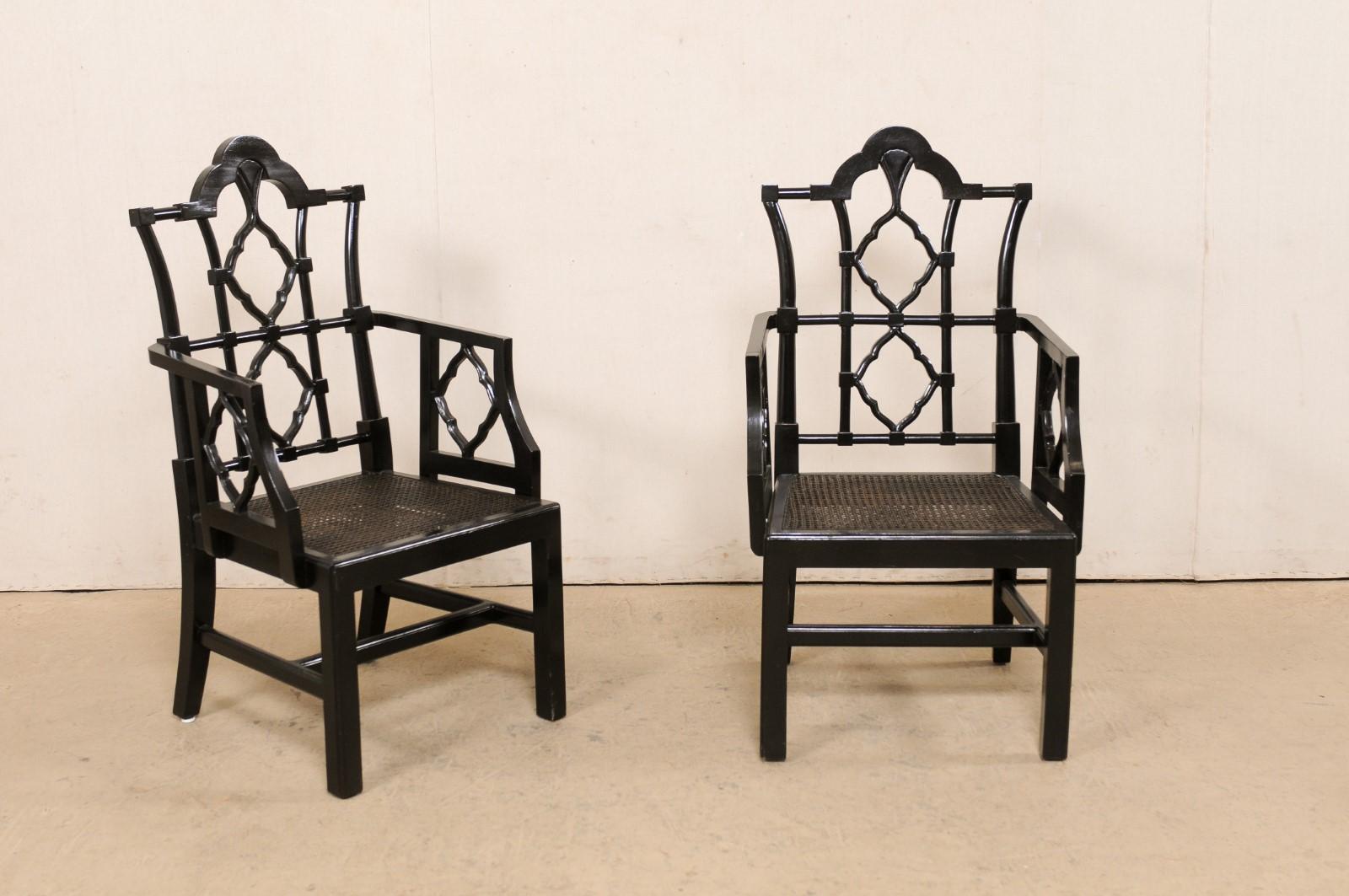 A pair of American-made wooden armchairs with cane seats. This vintage pair of Chippendale inspired occasional chairs have black wood frames, with raised-crest at back top-rails and a beautiful open lattice design, and hand-woven cane makes up the
