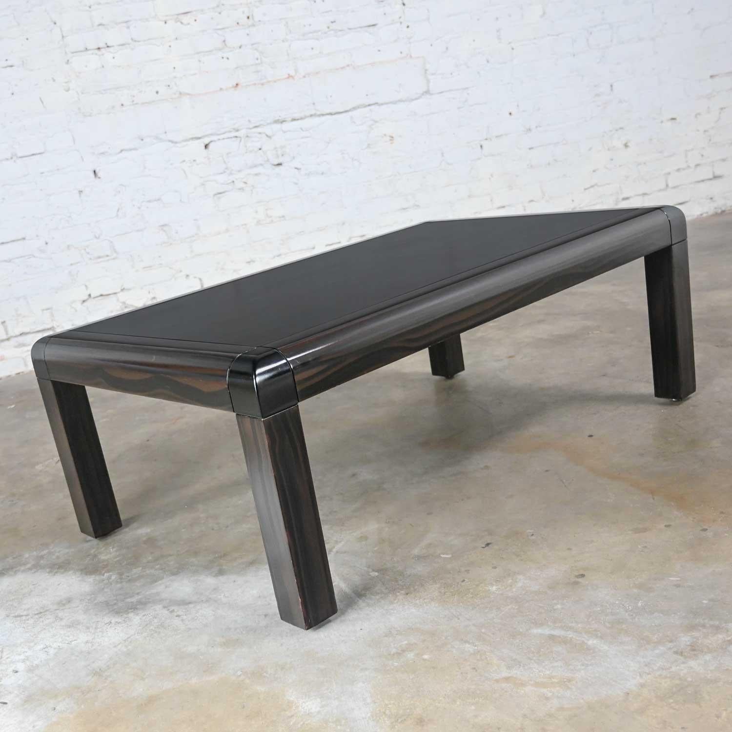 Handsome vintage modern coffee table with dark faux wood lacquered edges and legs, black lacquered waterfall style corners, and a black leather (possibly goatskin) top insert. The bottom of the table bears the prestigious leather tag signed by Karl
