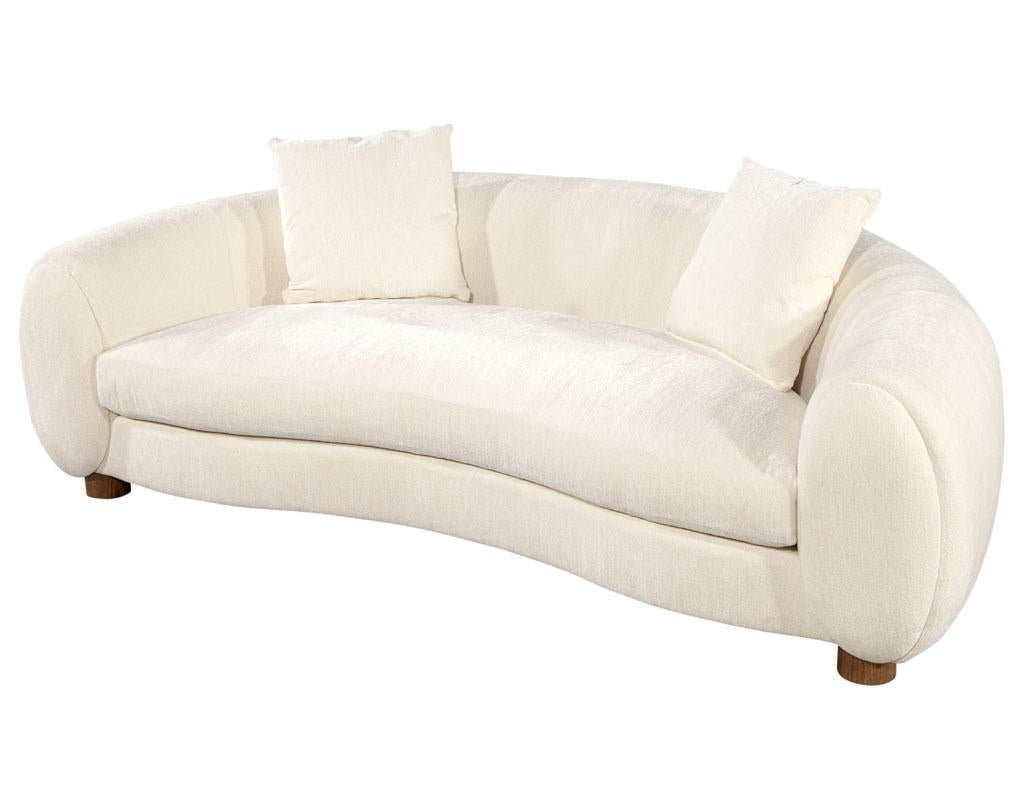 Modern curved linen sofa by Ellen Degeneres Perkins Sofa. Sophisticated curved design with clean lines. Sleek cylindrical feet in a light brown oak matte finish. Upholstered in a luxurious off-white fabric. 2 accent pillows included. Matching swivel
