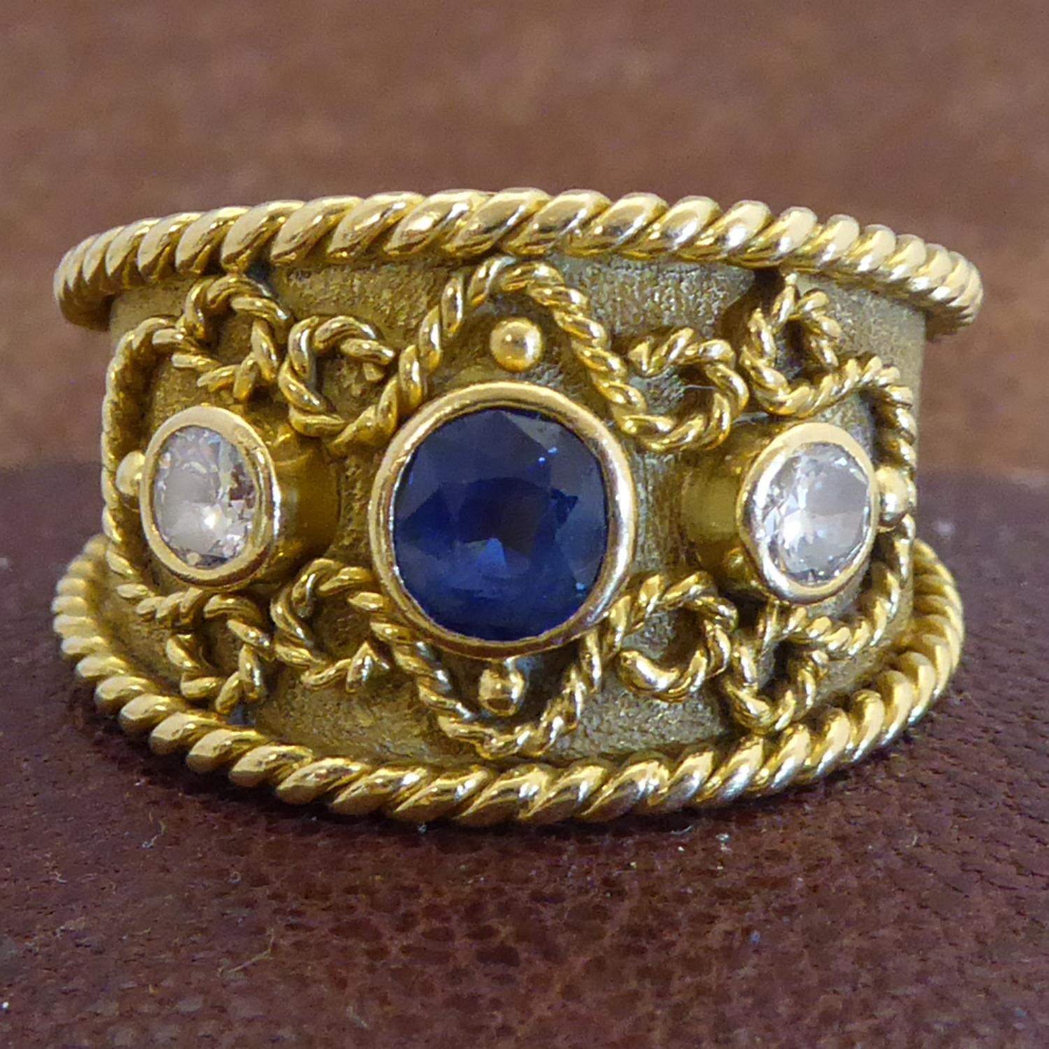 A modern sapphire and diamond Byzantine style fancy band ring.  Centrally set with a round mixed cut sapphire a round brilliant cut diamond to each side.  Rub over set to a yellow gold band with rope twist edges and a twisted wirework decoration