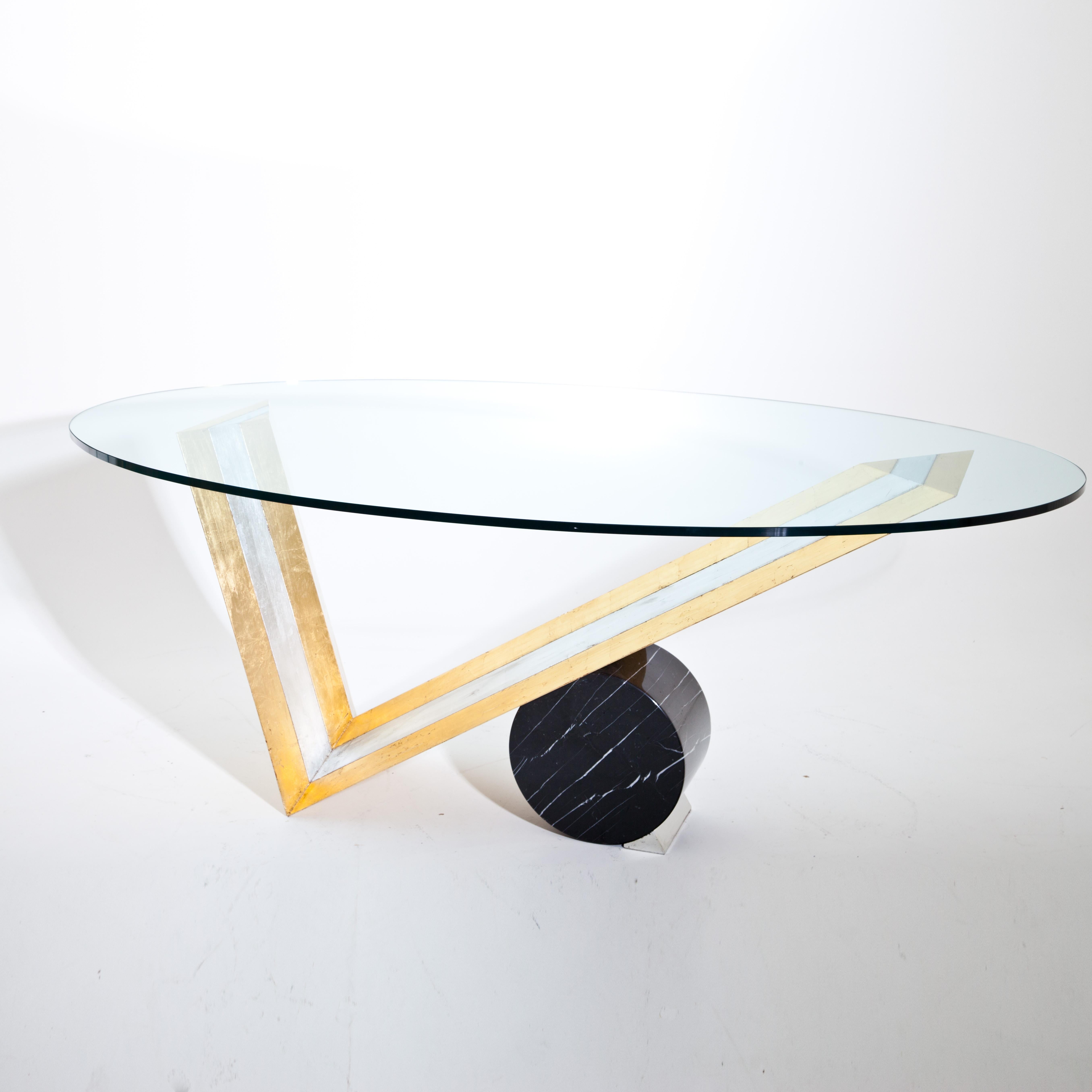 Large table with oval glass top on a V-shaped wooden frame with a large black marble cylinder block. The wooden frame is patinated in silver and gold.