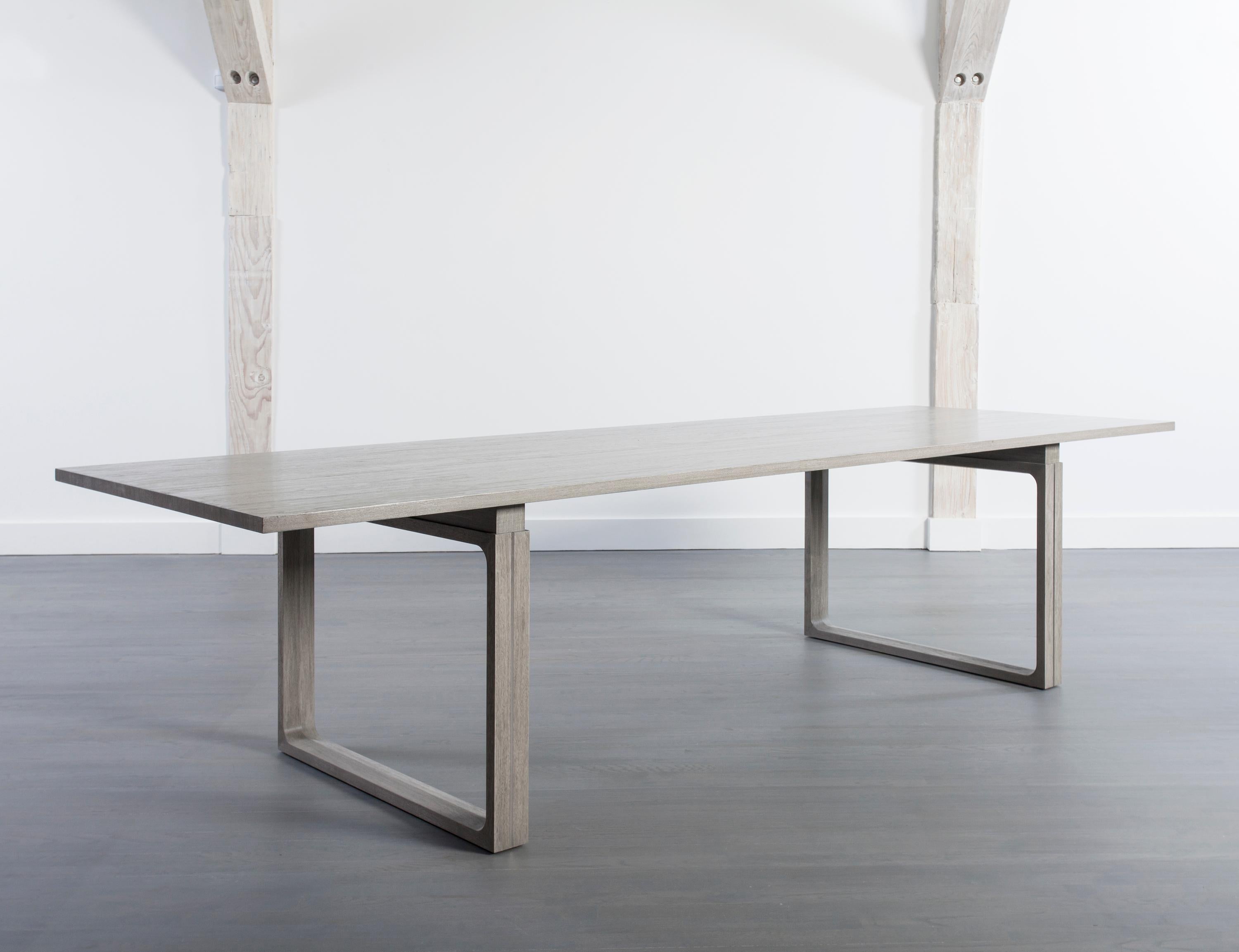 The clean lines of this elegant modern walnut dining table enables its wood grain to take center stage. Stained in a dove grey, and resting on a pair of sculptural open square legs, this graceful table will provide for both casual and elegant