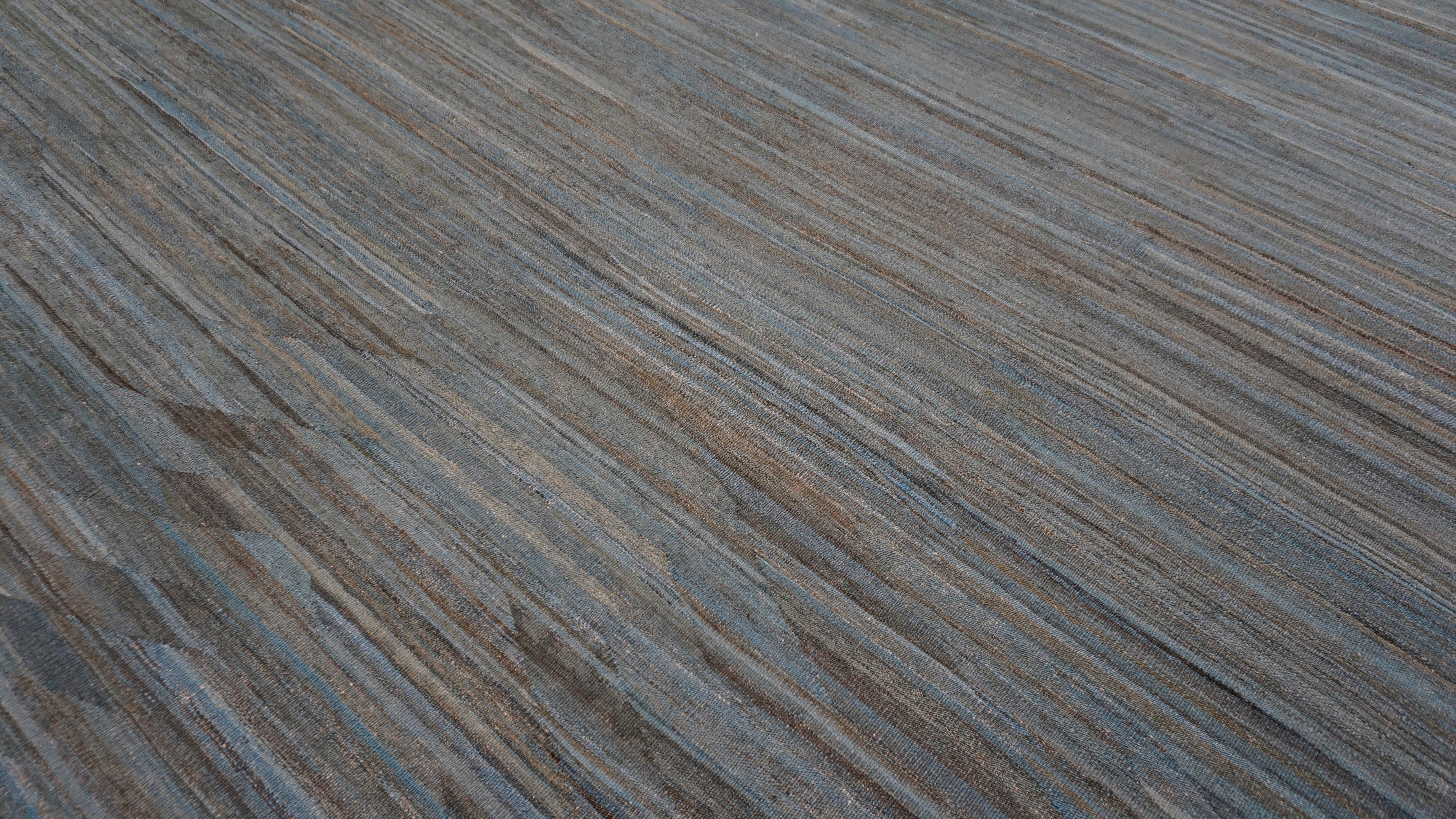 This handwoven wool kilim embodies a modern aesthetic rooted in traditional textile artistry. The color palette blends layers of blue, brown, and soft taupe, evoking the look of water-worn stone or aged denim. Featuring a subtle striated pattern