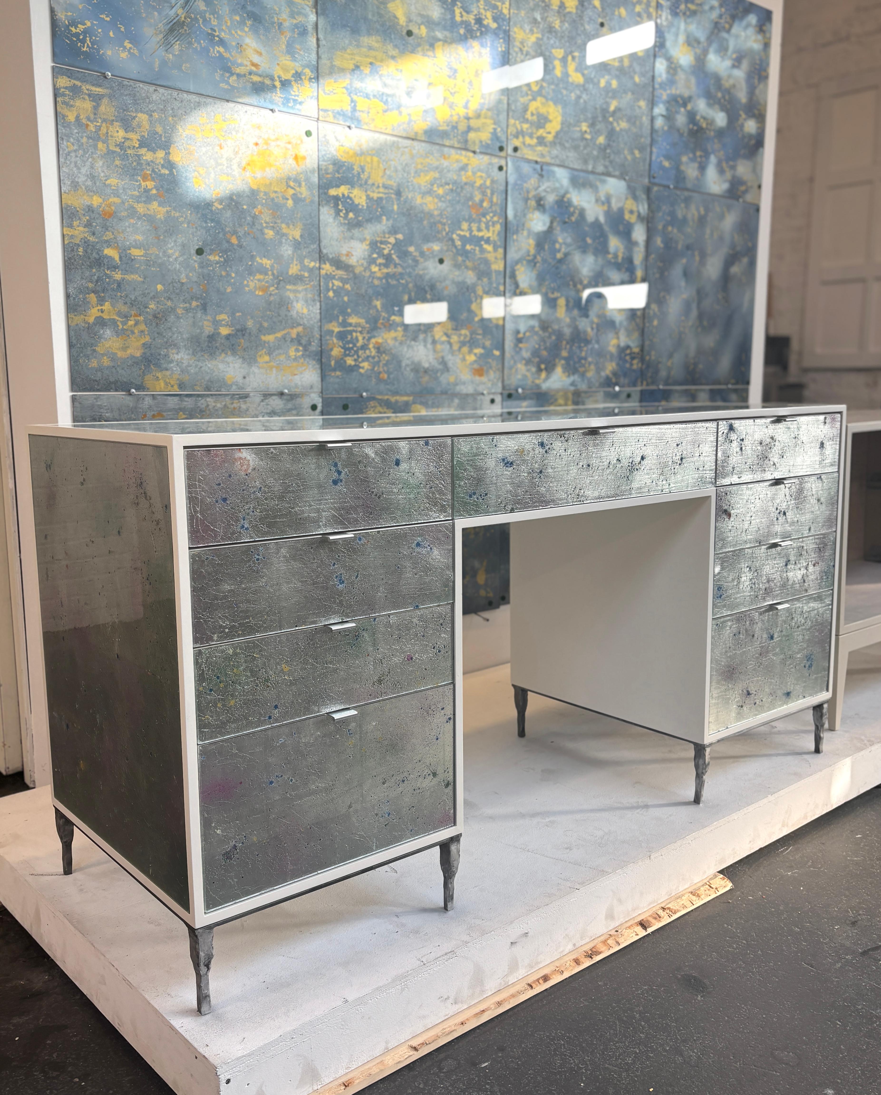 The Modern Mystic Lily Pond vanity by Ercole Home has a 9-drawer, with ivory wood finish on oak. Hand painted eglomise glass panels are inset on the surface. There are nine pulls in silver finish. The hand hammered metal base is in silver finish.