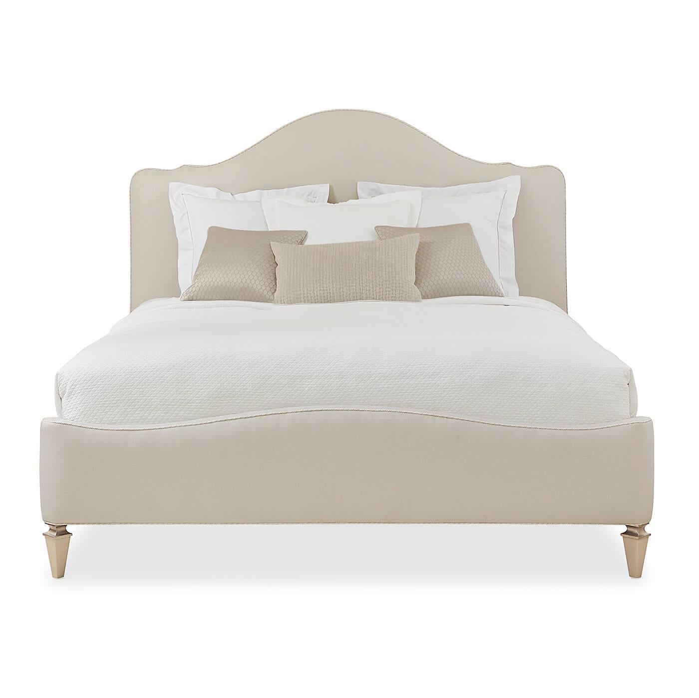 A Modern English camel-back king bed with exposed wood square tapered legs in rich tones of Sparkling Argent. This glamorous bed's headboard and footboard are upholstered in luxurious fabric and detailed with decorative cord trim.