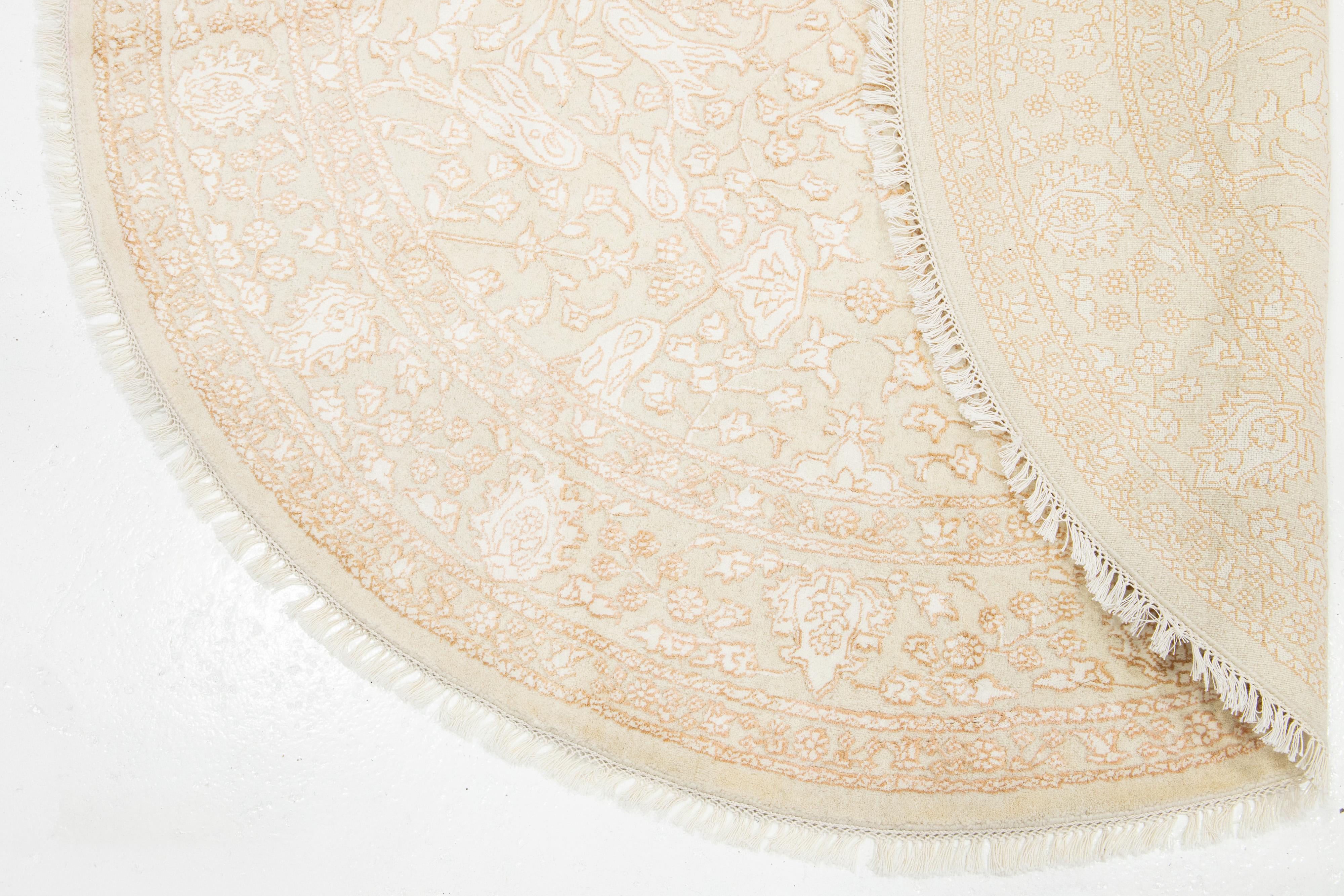 Dieser schöne, runde Teppich ist aus einer Mischung aus Wolle und Seide handgeknüpft und besticht durch seine beige Farbpalette. Mit eleganten Elfenbein- und Kupferakzenten verleiht das florale All-Over-Design jedem Raum eine künstlerische