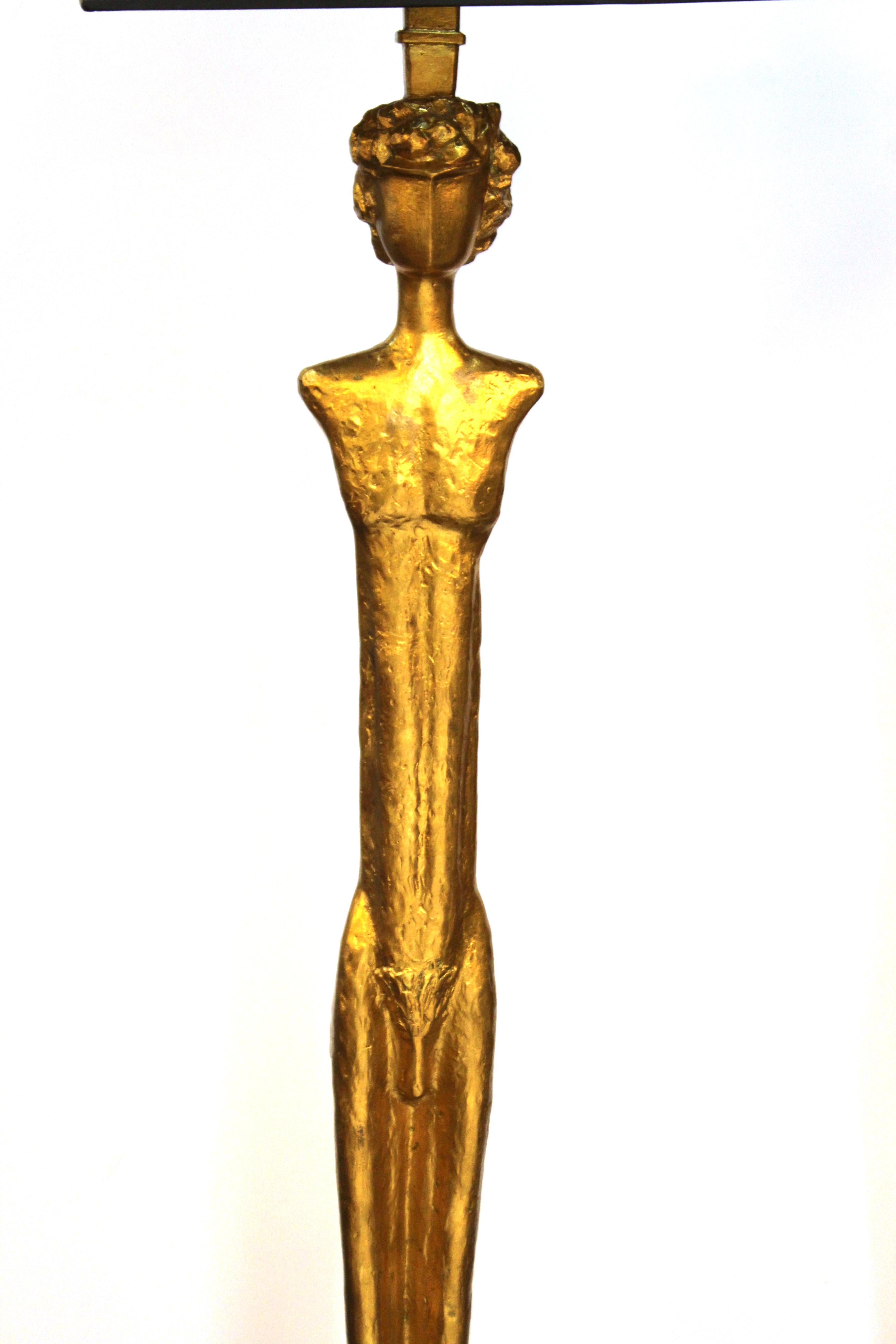 Modern pair of Giacometti style figural floor lamps in gilt bronze depicting elongated figures of a man in the nude and a woman in a dress, atop shallow stepped pedestals. The pair comes with black shades with a gilt interior, measuring 16 in. x 16