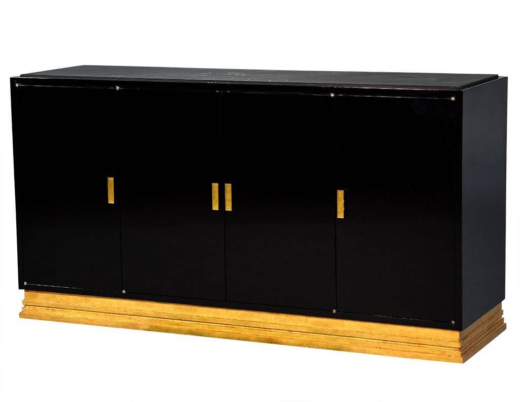 Modern gold leaf and black lacquer buffet console cabinet with stone top. This modern console is done in a high gloss black lacquer, topped with a black stone insert and sits atop a gold leafed pedestal base. There are two sleek doors at each end