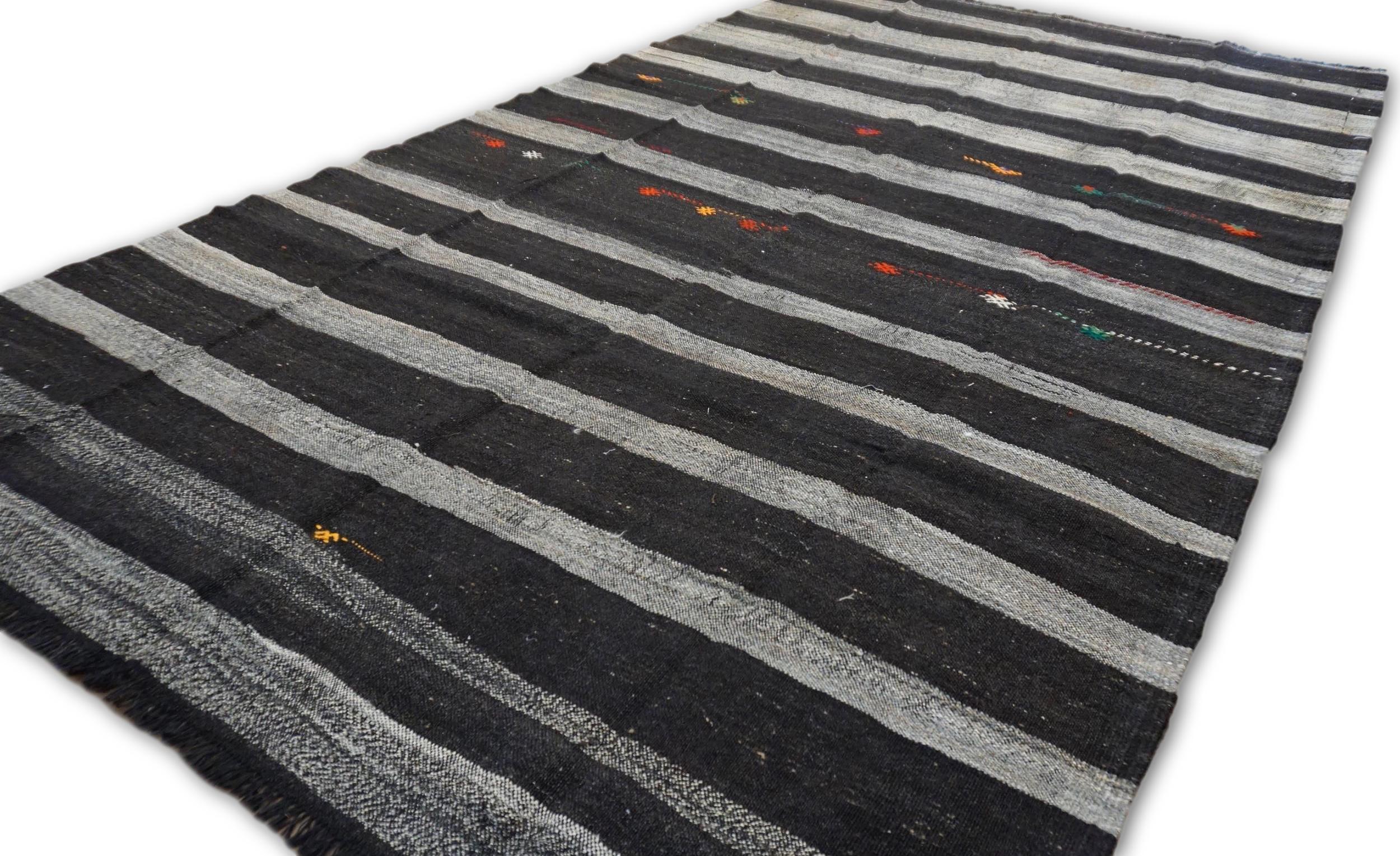 Questo Kilim vintage in pelo di capra presenta un sorprendente motivo a strisce, sottilmente arricchito da delicati e colorati ricami tribali. Il contrasto tra le audaci strisce monocromatiche e i vivaci accenti decorativi rende questo pezzo una