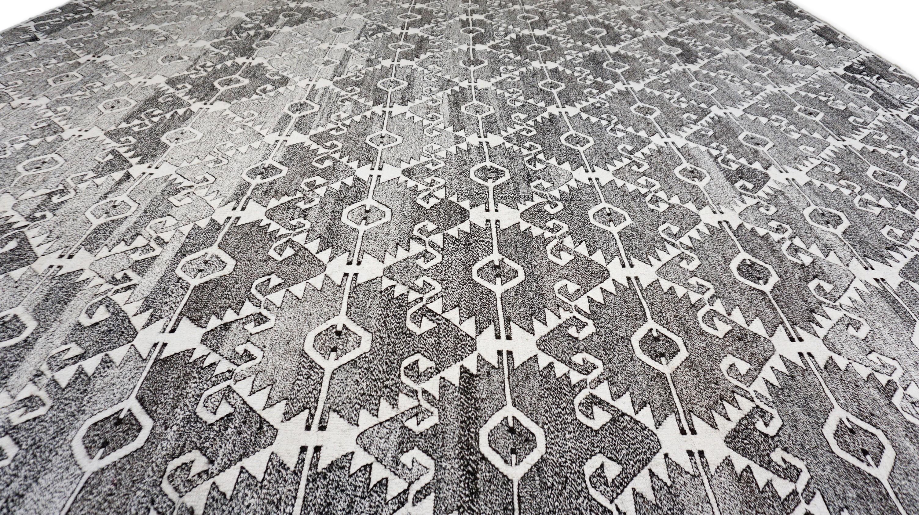 This hand-woven wool rug showcases a bold and captivating geometric tribal design, rendered in a refined monochrome palette of charcoal, grey, and white. Expertly crafted by skilled artisans, this rug features repeating angular motifs inspired by