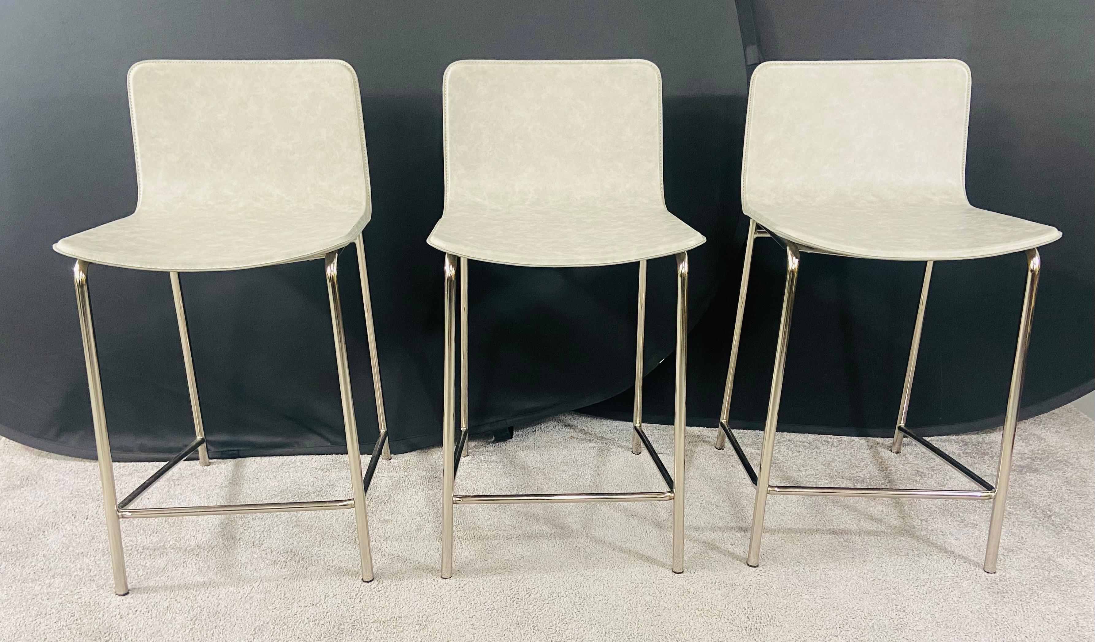 A set of three modern quality bar or kitchen stools. The stools seats are made of leather in a Grey neutral color and are well stitched. The frame of the stools is made in stainless steel. Modern, elegant and minimalist, the stools will elevate any