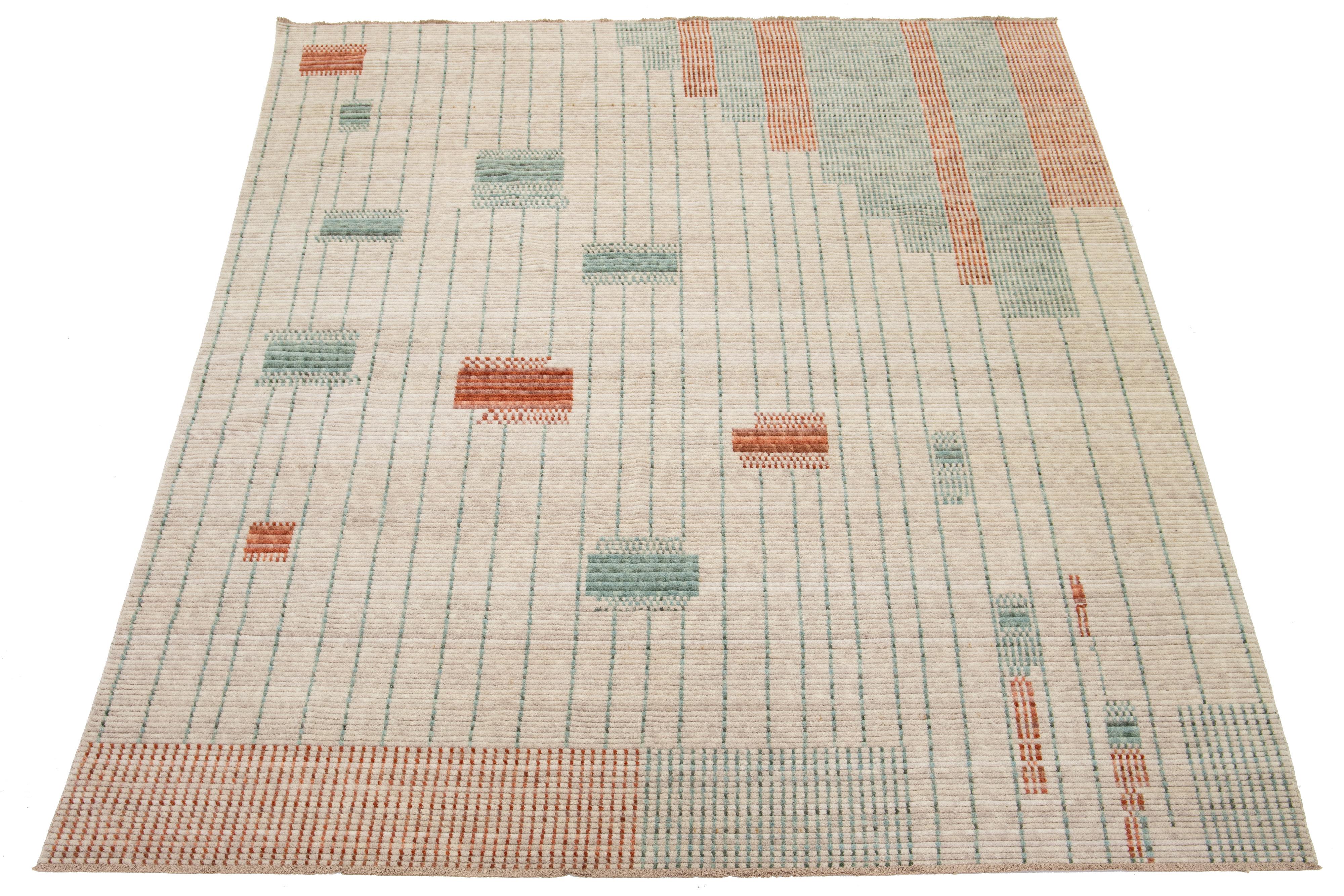 Dieser handgewebte moderne Wollteppich ist ein echter Hingucker. Der elegante, hellbeigefarbene Hintergrund ist die perfekte Leinwand für das verschlungene geometrische Muster in leuchtendem Orange und Grün, das jeden Raum optisch aufwertet.