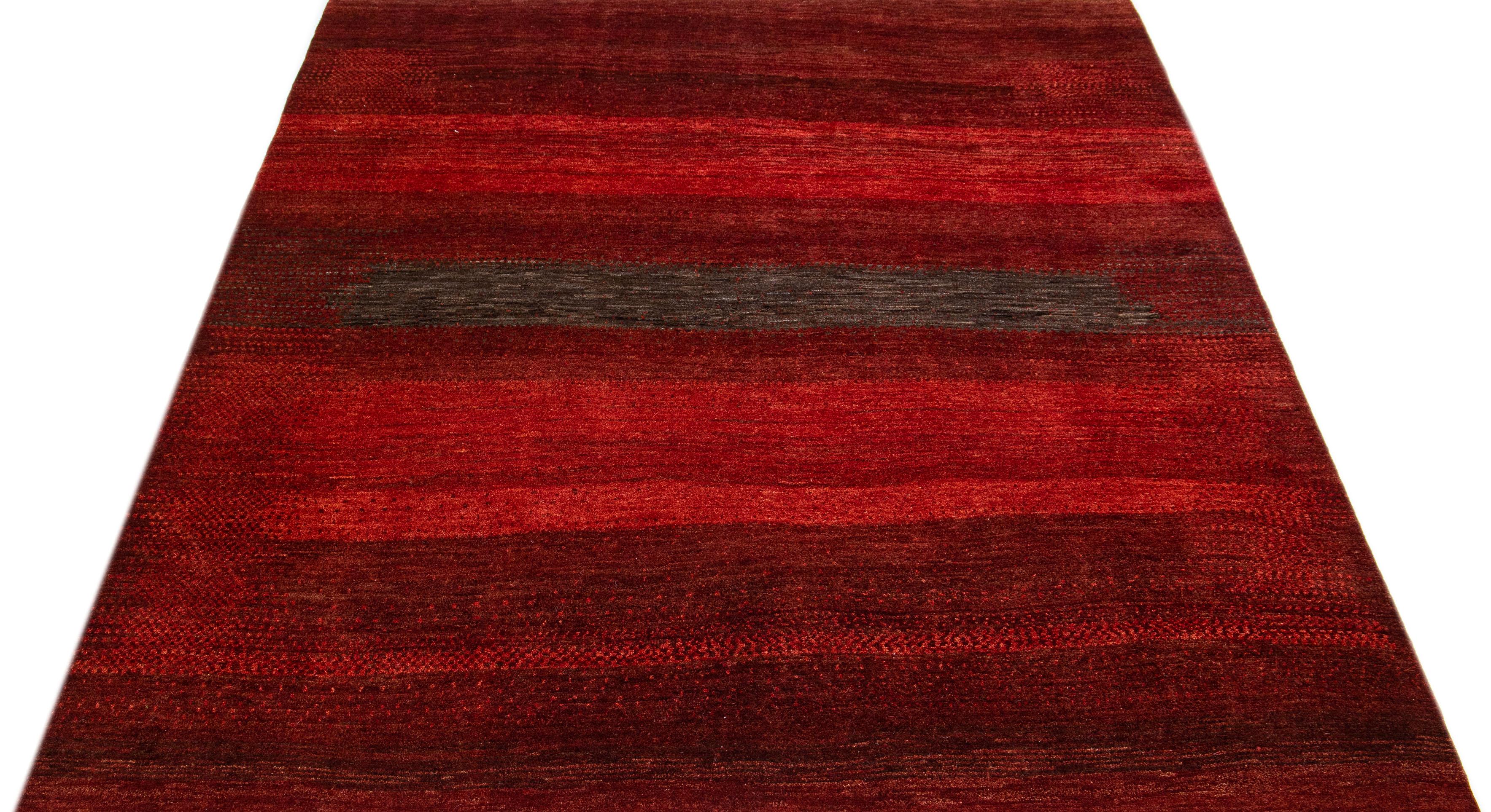 Dieser handgefertigte Wollteppich im Gabbeh-Stil zeigt eine abstrakte Ästhetik mit braunen Akzenten auf einem leuchtend roten Farbfeld.

Dieser Teppich misst 7'8