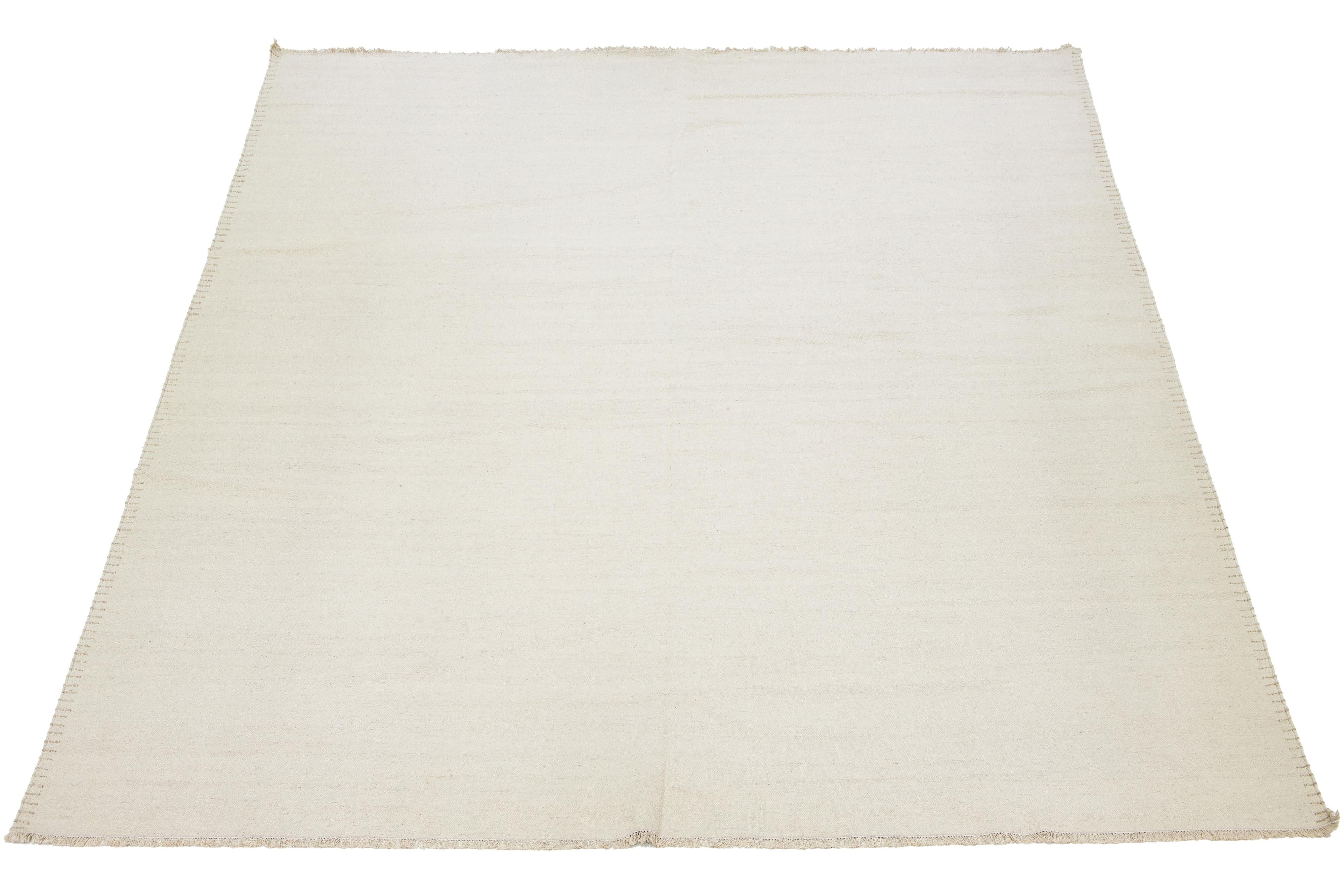 Dieser flachgewebte Teppich zeigt ein modernes Dhurrie-Design mit einer beigen Grundfarbe und dekorativen Akzenten entlang der Bordüre.

 Dieser Teppich misst 12'5