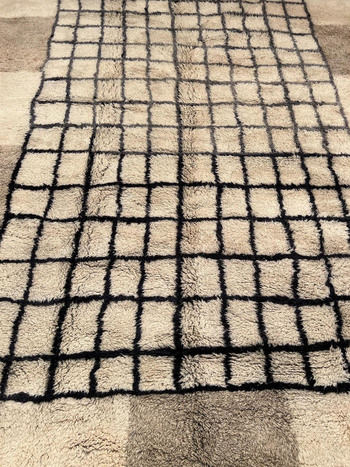 This graphic, handmade Moroccan rug features a unique geometric design on a natural wool base. This handwoven rug features a neutral cream wool base with an interesting, varied geometric grid pattern in black, grey and taupe wool. Handmade on a loom