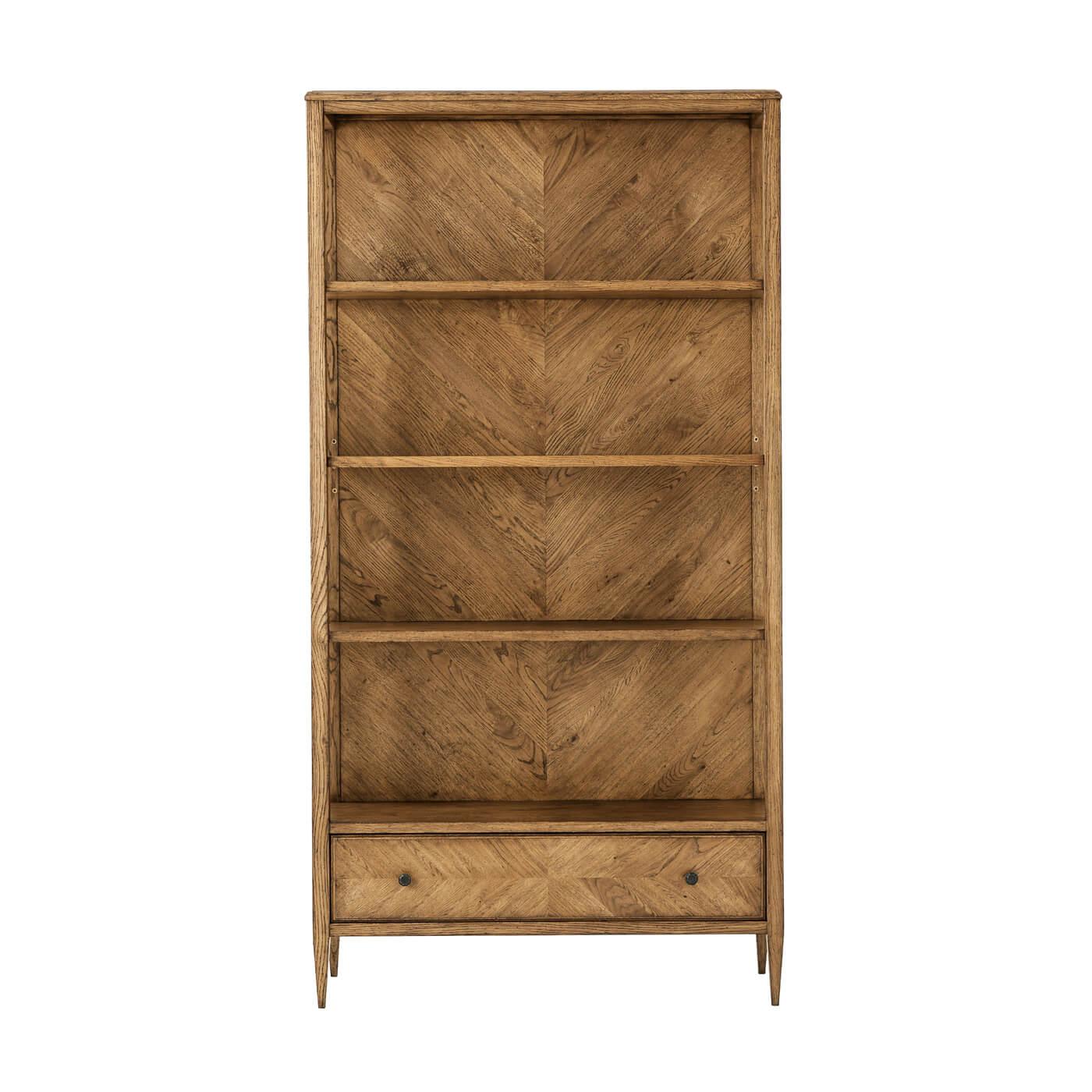 A Modern herringbone parquetry bookcase in our light oak Dusk finish with three veneered shelves and a herringbone parquetry design on the bottom drawer. It sits on tapered legs with Verde Bronze finished knobs. A beautiful functional piece for