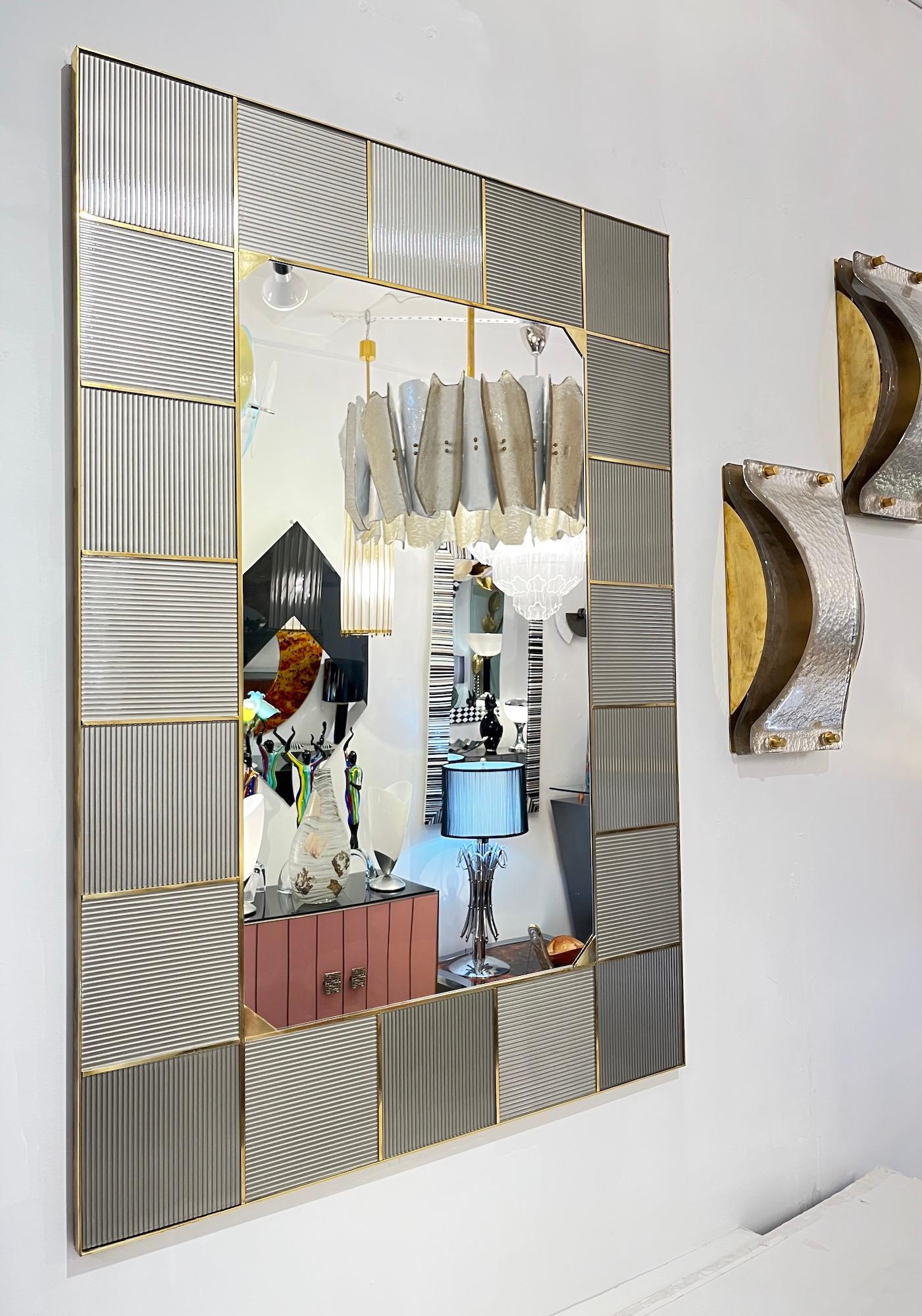 Este espejo contemporáneo italiano Couture Design de color gris piedra y latón tiene una construcción de alta calidad, con un marco que lleva incrustado un abrazador borde de latón en todo el contorno, así como en el borde interior de la placa