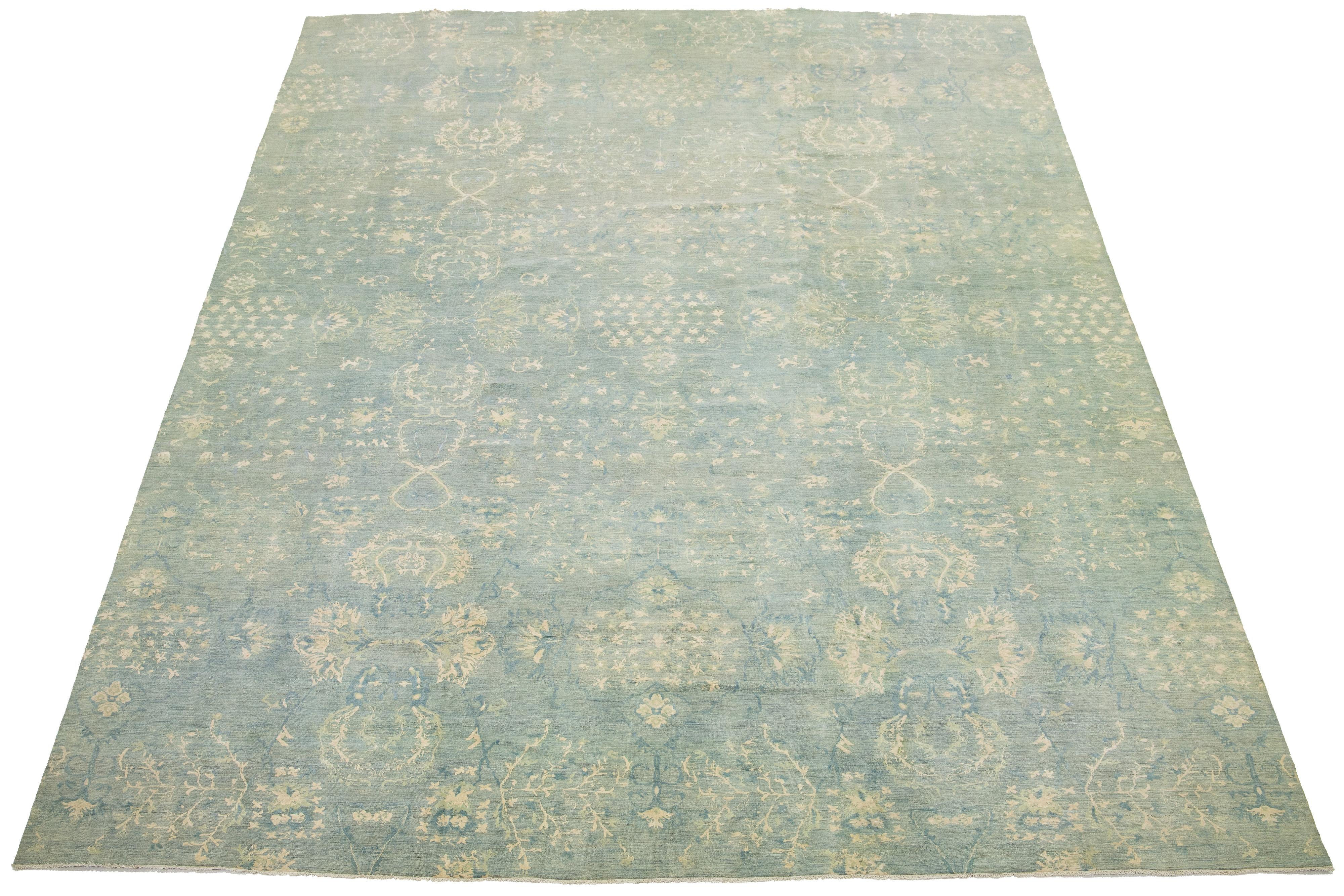 This is an oversized contemporary Khotan wool rug from India. The design showcases a deep blue and green base with intricate cream-colored patterns. These include eye-catching floral elements.

This rug measures 13'9