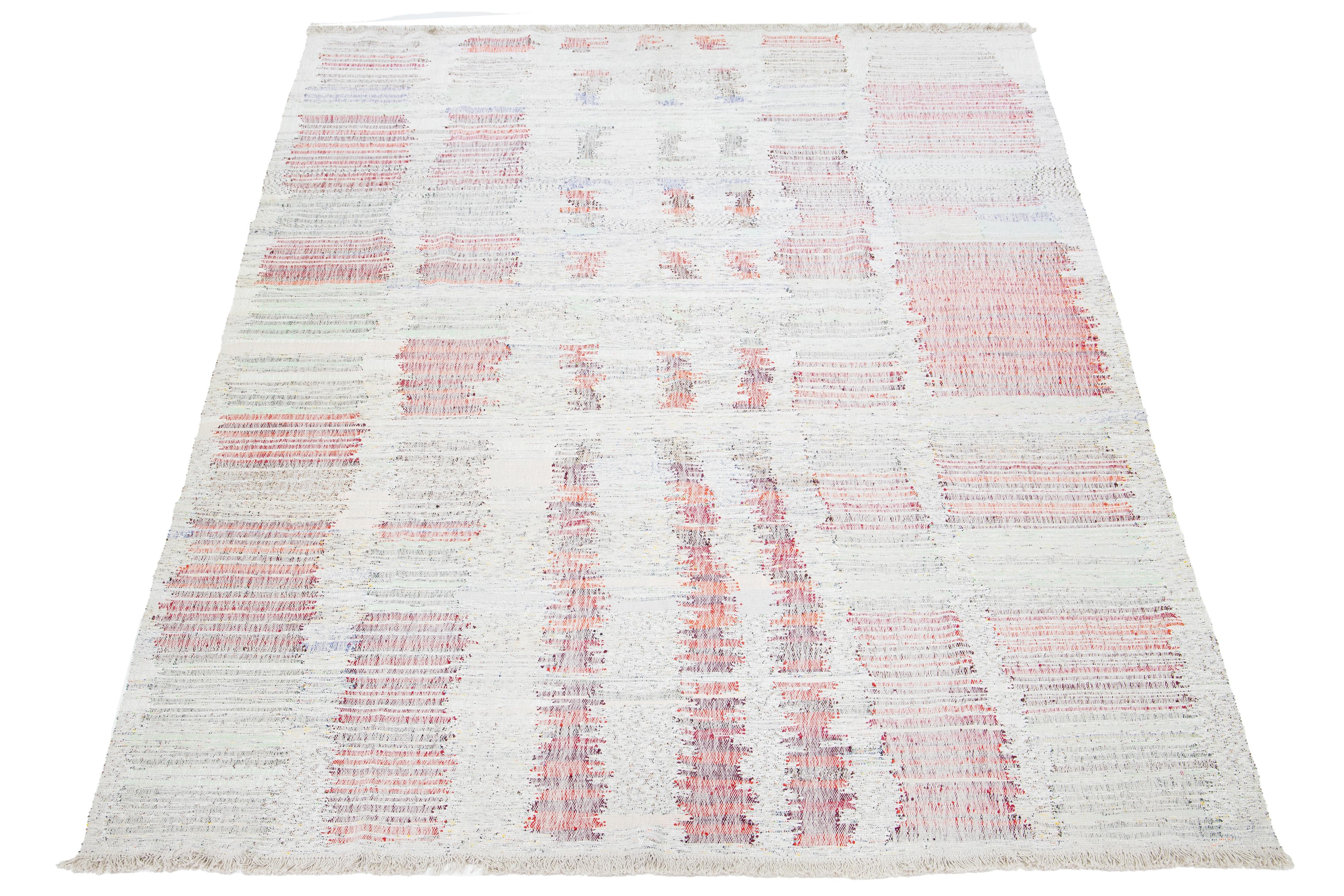 Dieser Teppich zeigt ein modernes Flachgewebe im Kilim-Stil, das aus Wolle gefertigt ist. Es hat ein beiges Feld mit einem mehrfarbigen Streifenmuster.

Dieser Teppich misst 9'6