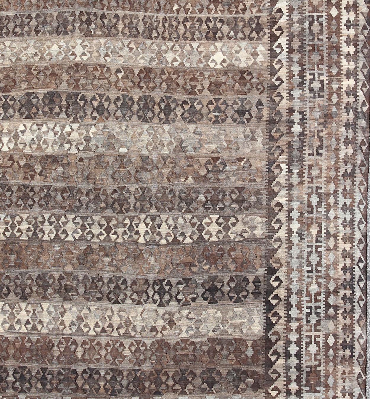 Cet exquis grand Kilim d'Afghanistan présente un motif tribal répétitif avec des rayures alternées dans différentes couleurs naturelles au centre, dans différentes nuances de brun, de gris et de blanc, entouré d'une bordure de motifs étonnants dans