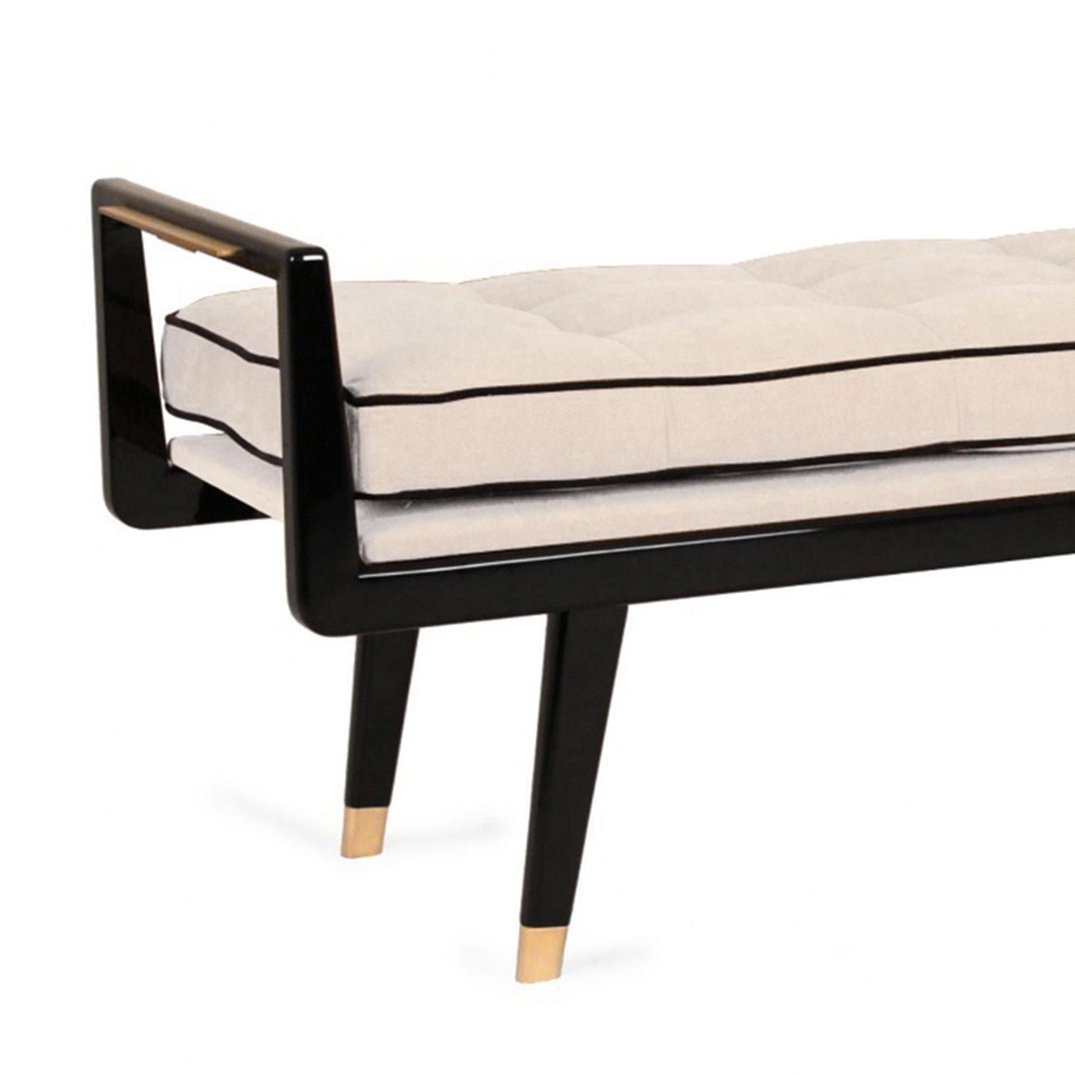 Clean-lined bench with angular frame and midcentury styling. The lacquered body features tapered legs enhanced with brushed brass feet caps. Sleek brass details also adorn each end. The upholstered seat is topped with a button tufted cushion with