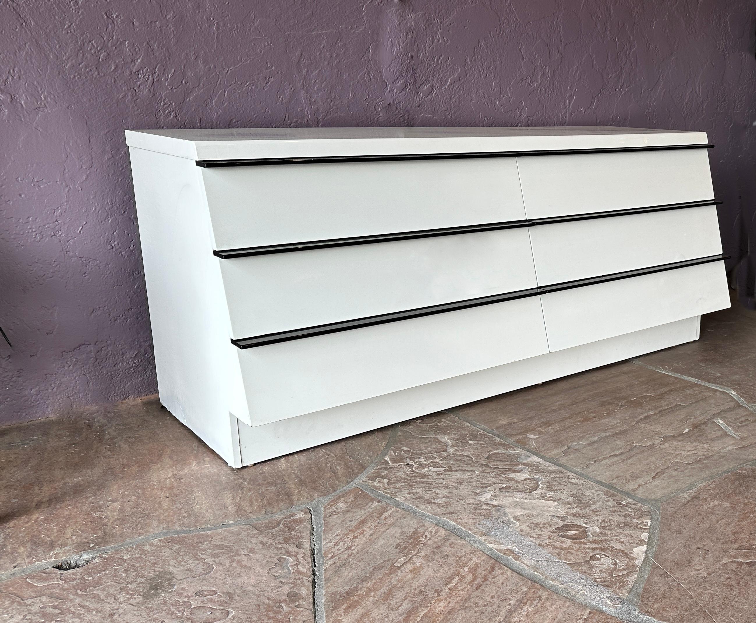 This white lacquered dresser  from the 1980s, reminiscent of the Roger Rougier style, boasts a unique feature: contrasting black handle accents adorning six stairs-stepped drawers .

With its refined high-gloss finish, the dresser's appeal lies in